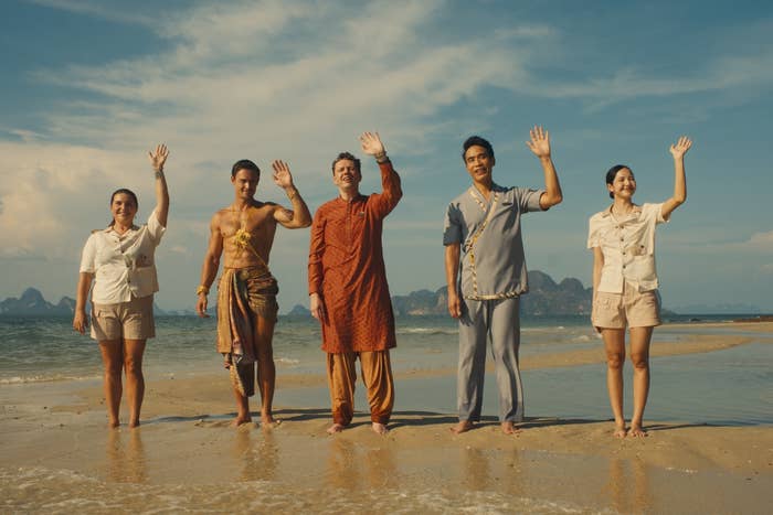 Five people stand on a beach, waving. They wear a mix of casual and traditional outfits, with a scenic ocean and mountain backdrop