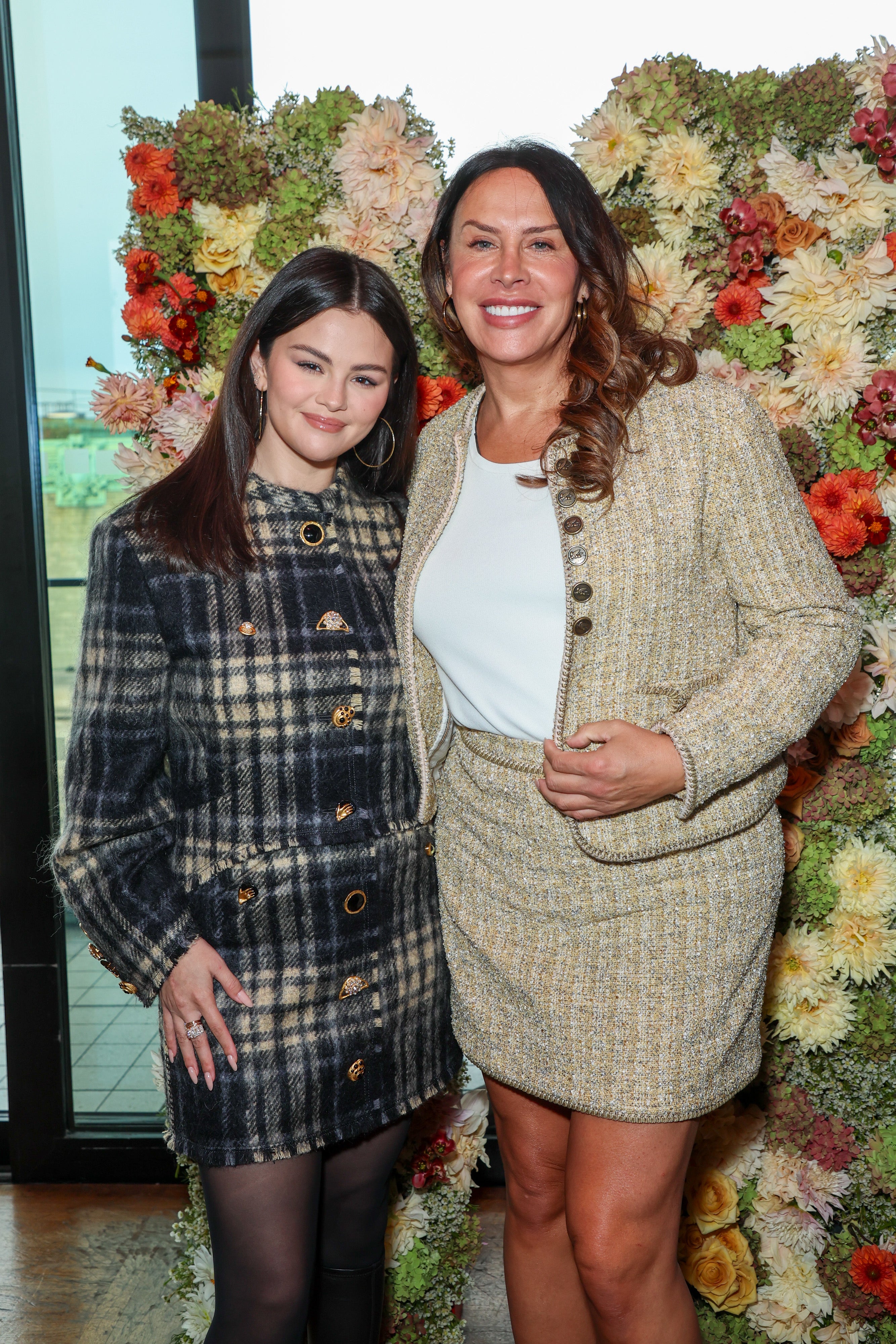 Selena Gomez and Karla Sofia Gascon smiling in front of a floral backdrop. One is wearing a plaid coat, and the other a tweed skirt suit
