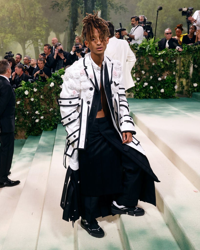 Individual in a long, patterned coat and layered clothing poses on an elegant staircase at a formal event