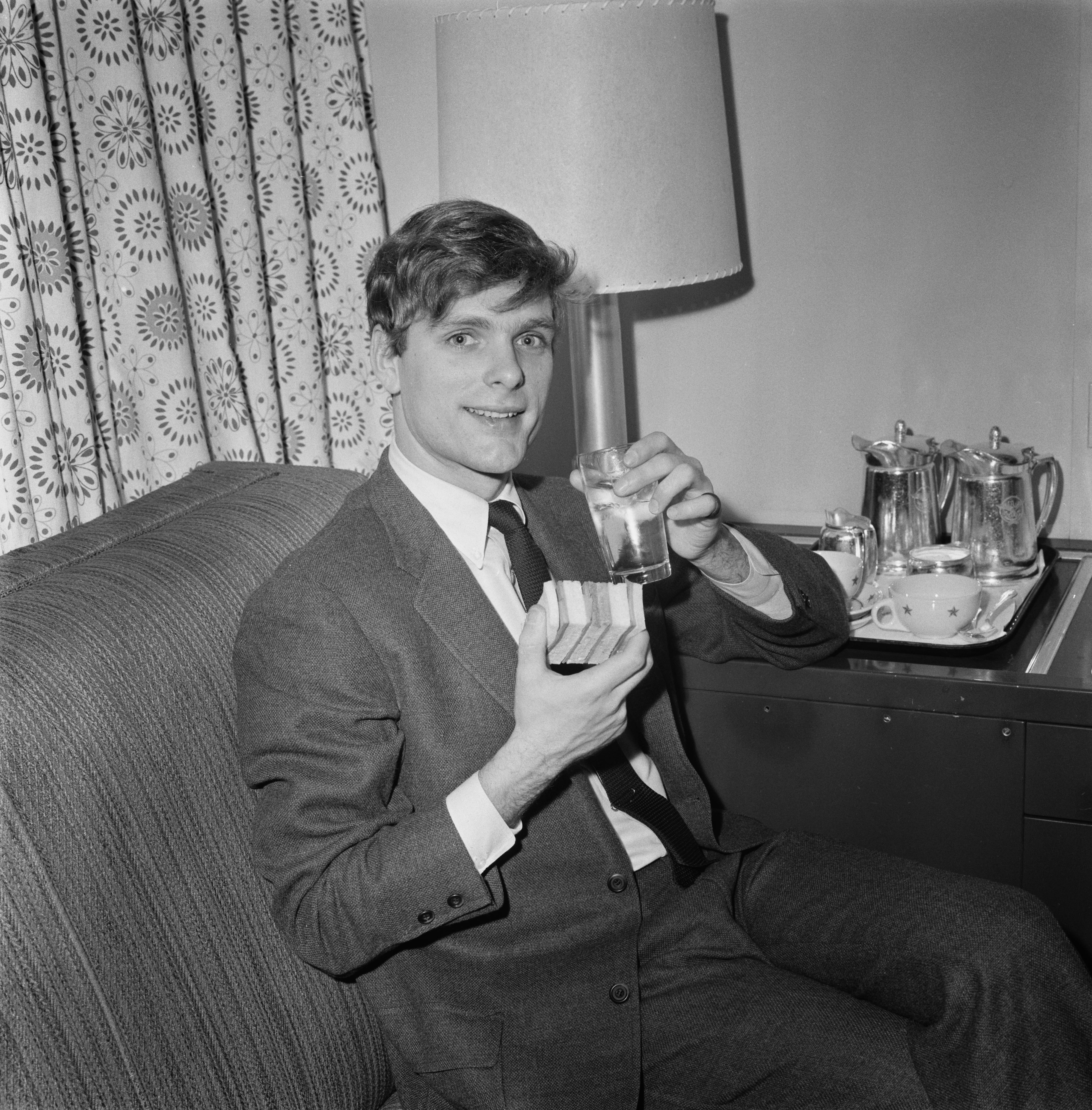 Person in a suit holding a drink and a box, sitting on a couch in a room with a lamp and a small table set with a tea service