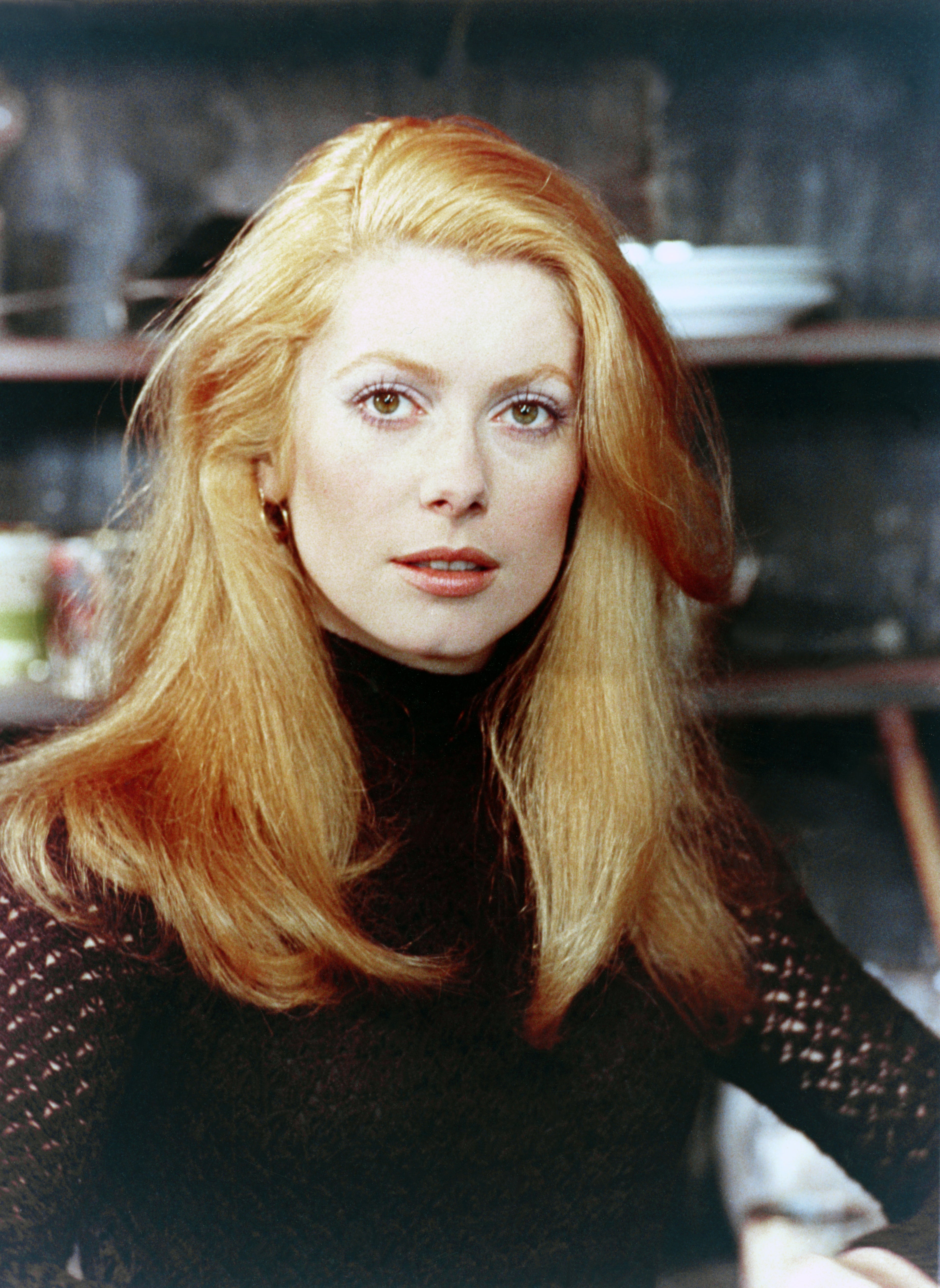 Person with long hair, wearing a textured turtleneck sweater, stands with a neutral expression indoors
