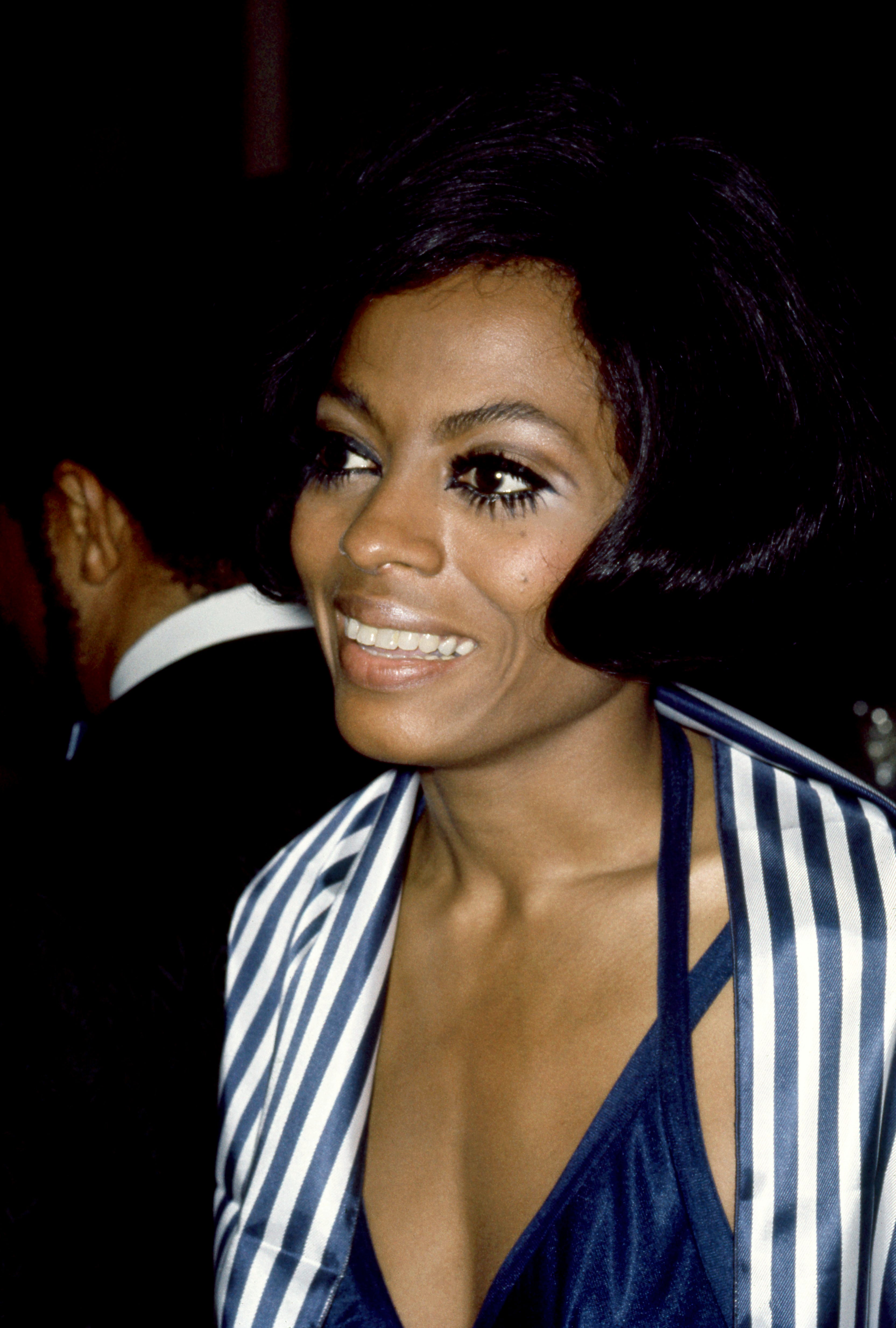 A person smiling, wearing a stylish striped jacket over a V-neck top, with bold eye makeup, on an event background