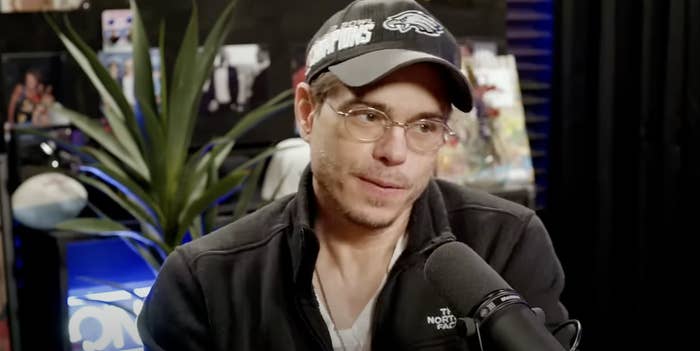 Matthew Lawrence in casual attire and cap speaking into a microphone, with a plant and various items in the background