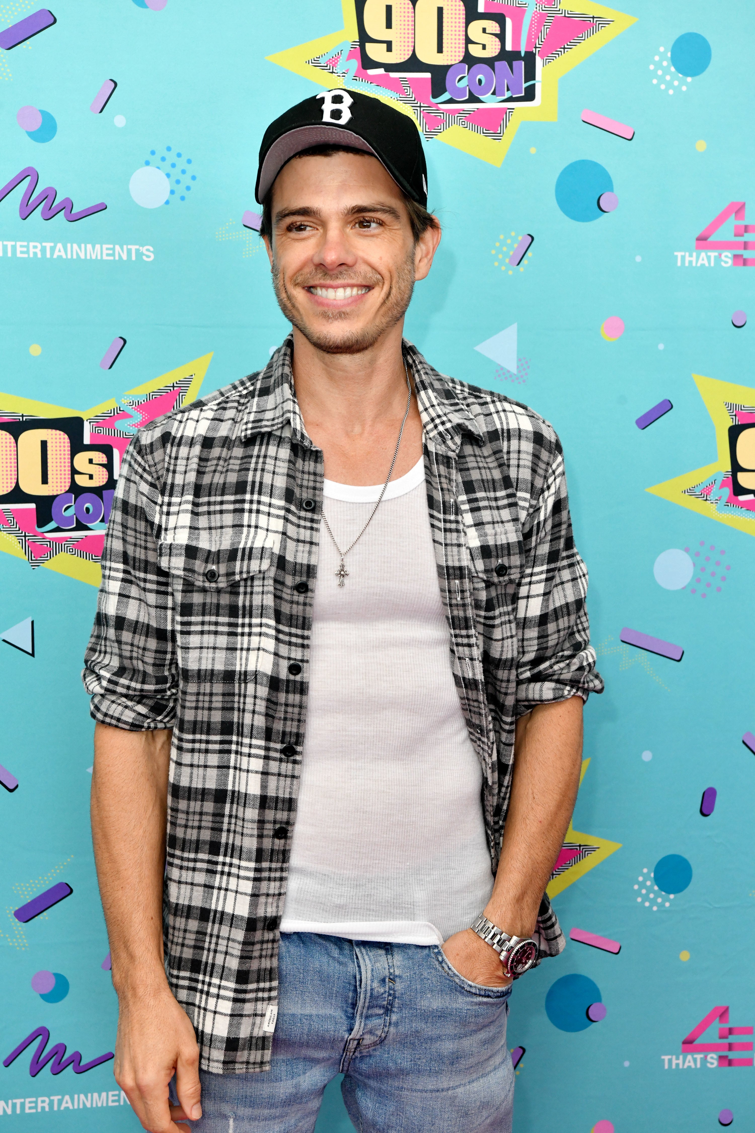 Matthew Lawrence in casual attire with a plaid shirt, tank top, and baseball cap, standing in front of a '90s-themed backdrop