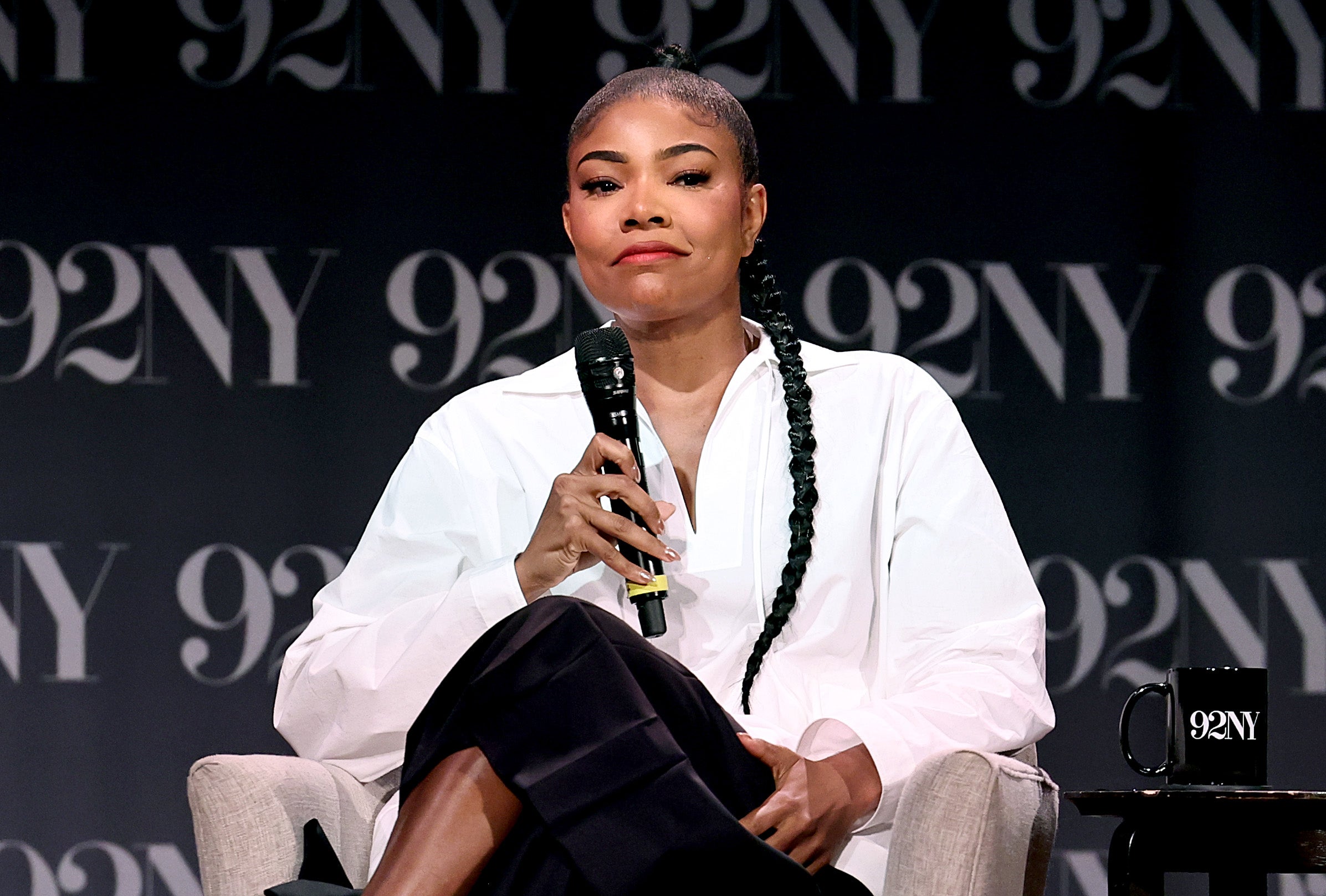 Gabrielle Union with long braid in white shirt, holding a microphone, seated at an event with "92NY" backdrop