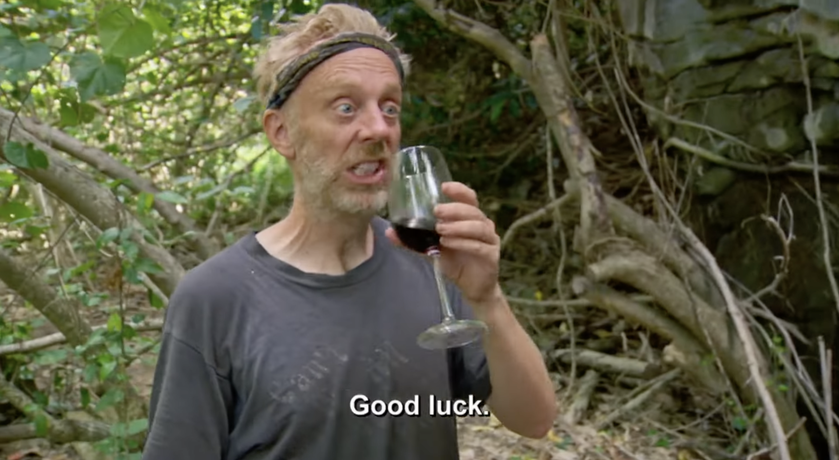 A person with a headband and casual shirt holds a wine glass, standing in a jungle setting. The caption reads, "Good luck."