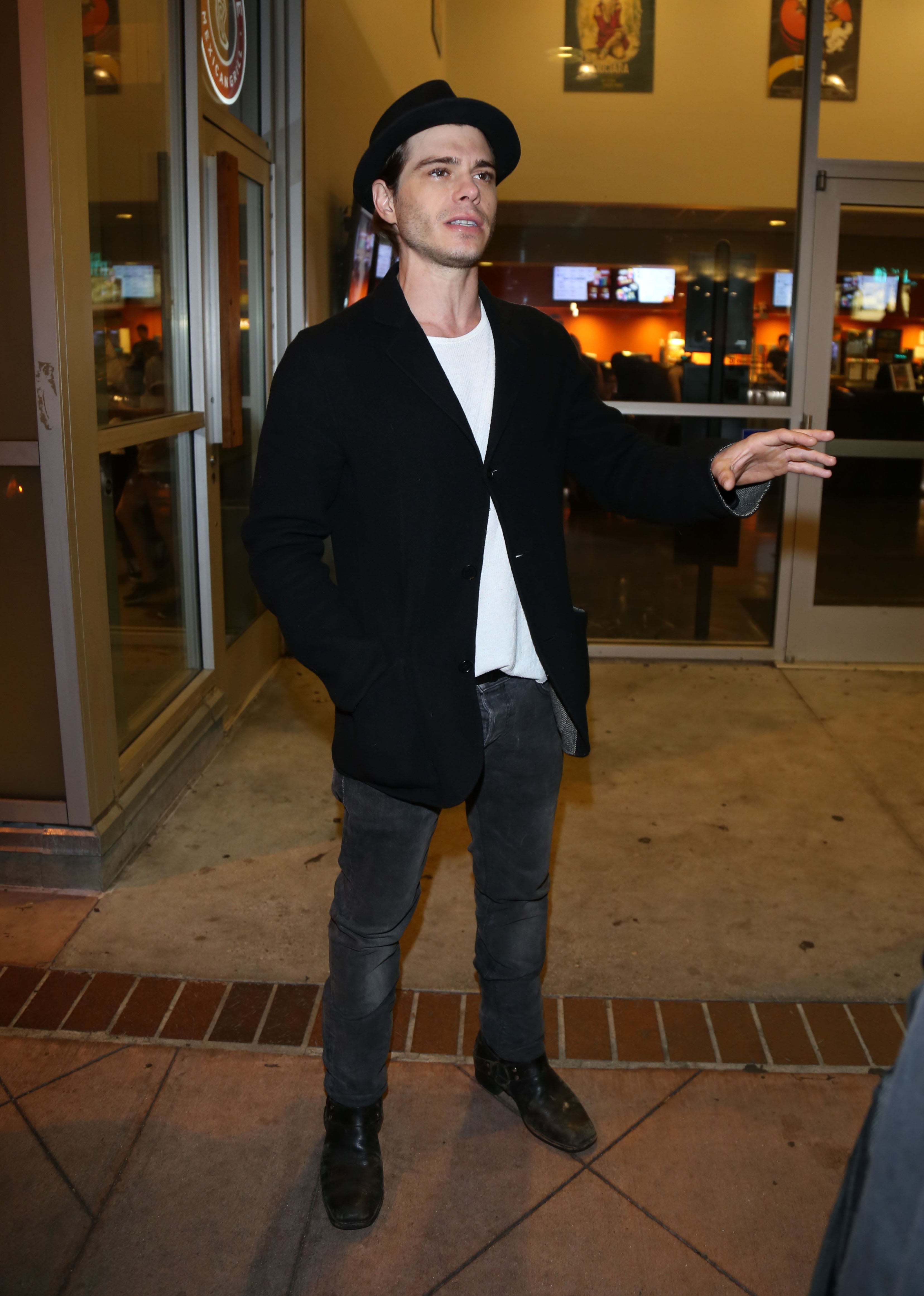 Matthew Lawrence in a jacket, shirt, and jeans is standing outside a building, gesturing with one hand