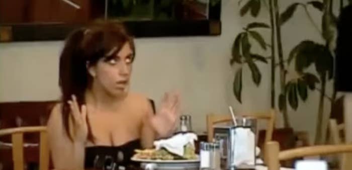 A woman sits at a restaurant table with a plate of food, appearing surprised or animated during a conversation