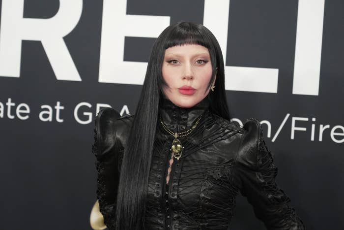 Person in a sleek, black leather outfit with long dark hair, attending a formal event in front of a backdrop with text