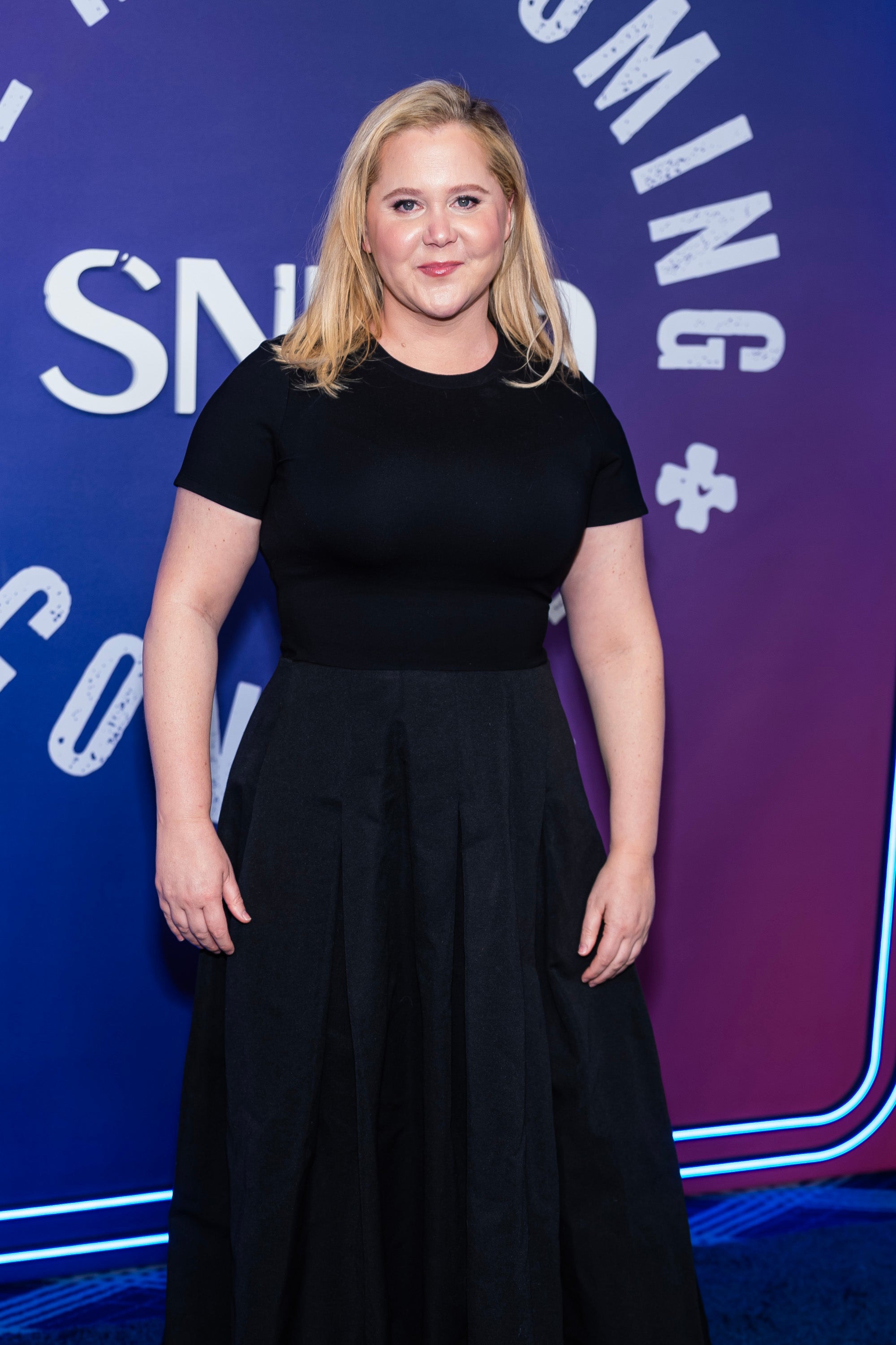 Person in a black top and flowing skirt at an event with a patterned backdrop