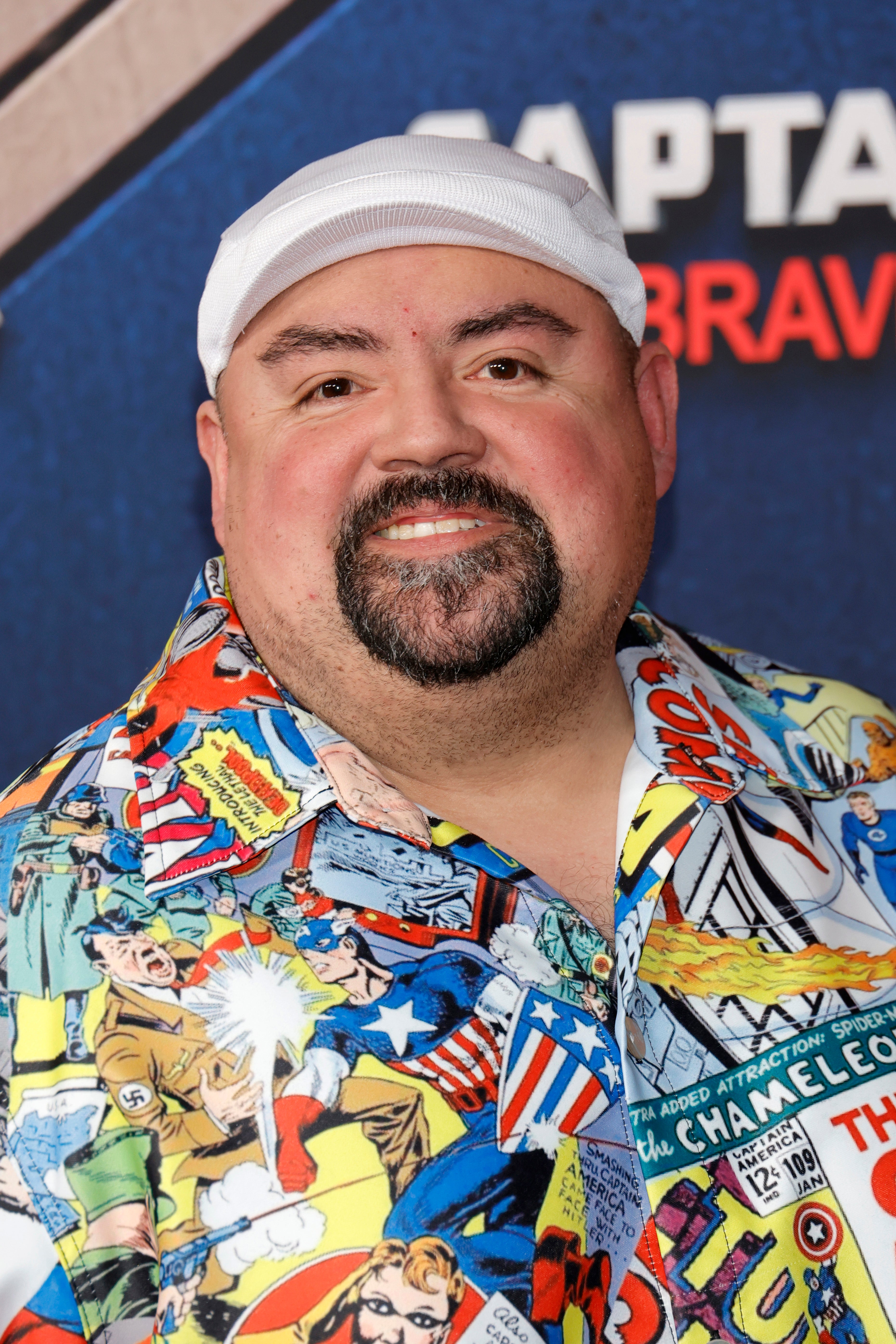 Man in a comic-themed shirt and white cap smiles at a red carpet event
