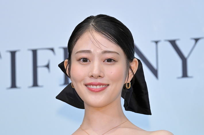 女性が黒いオフショルダードレスを着て、笑顔でポーズ。背景にブランド名。