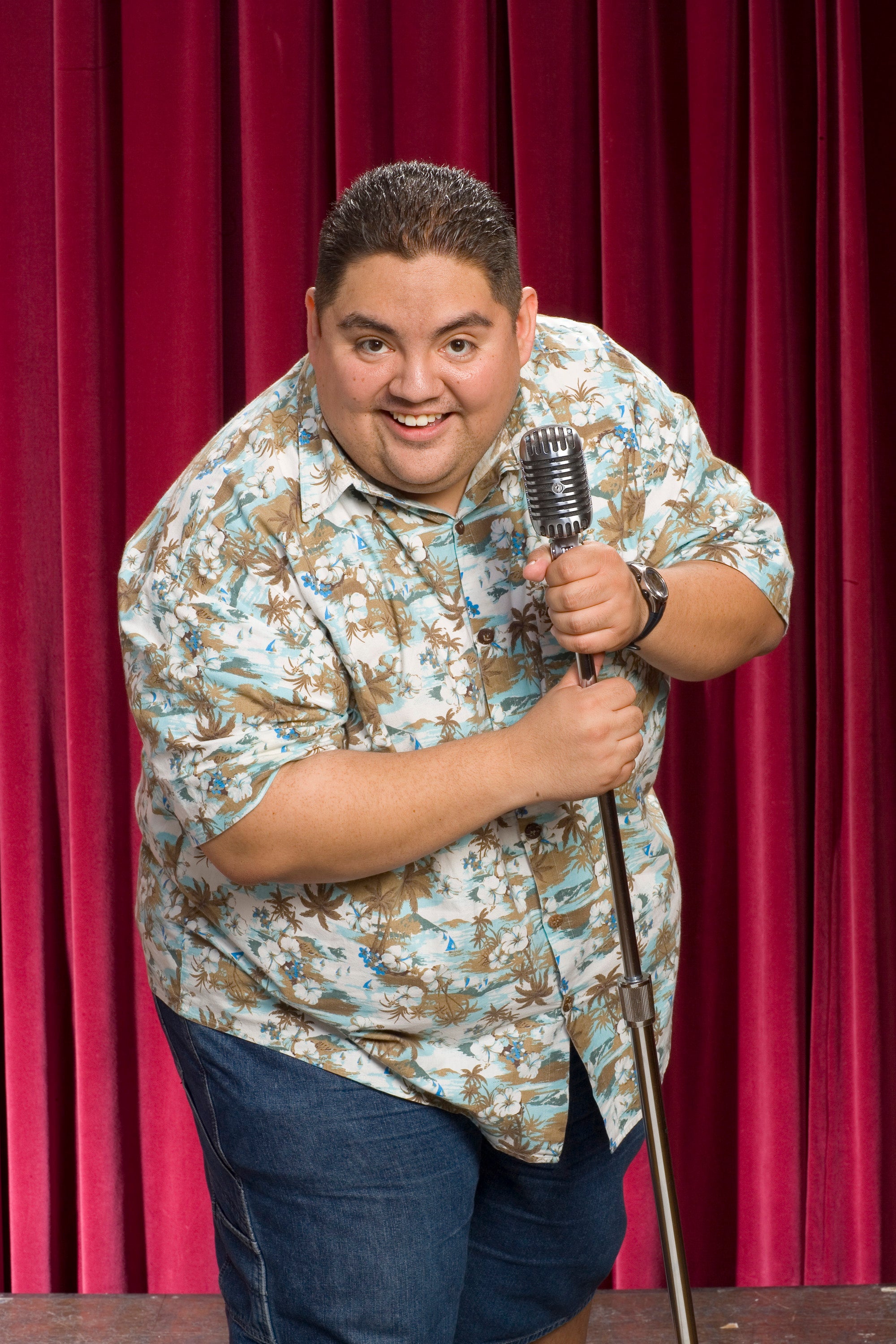 Person holding a microphone, smiling, wearing a floral shirt and denim pants, standing in front of red curtains