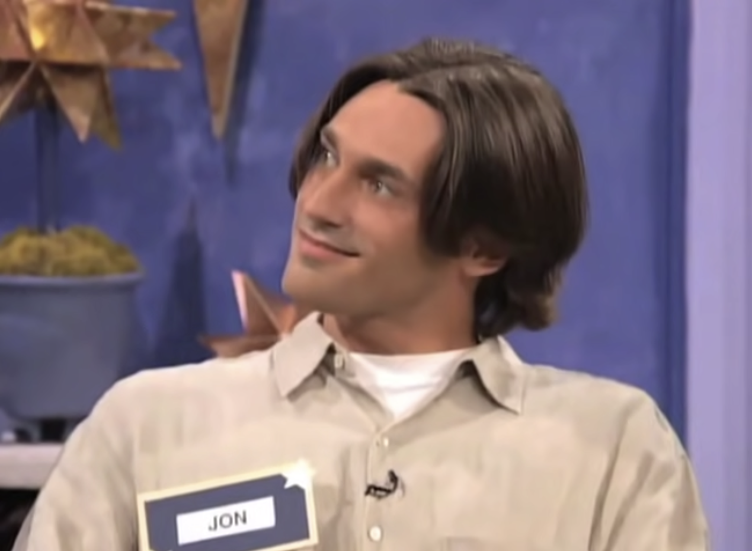 Person with medium-length hair, wearing a casual button-up shirt, looks to the side smiling, seated in an indoor setting with decorative elements in the background