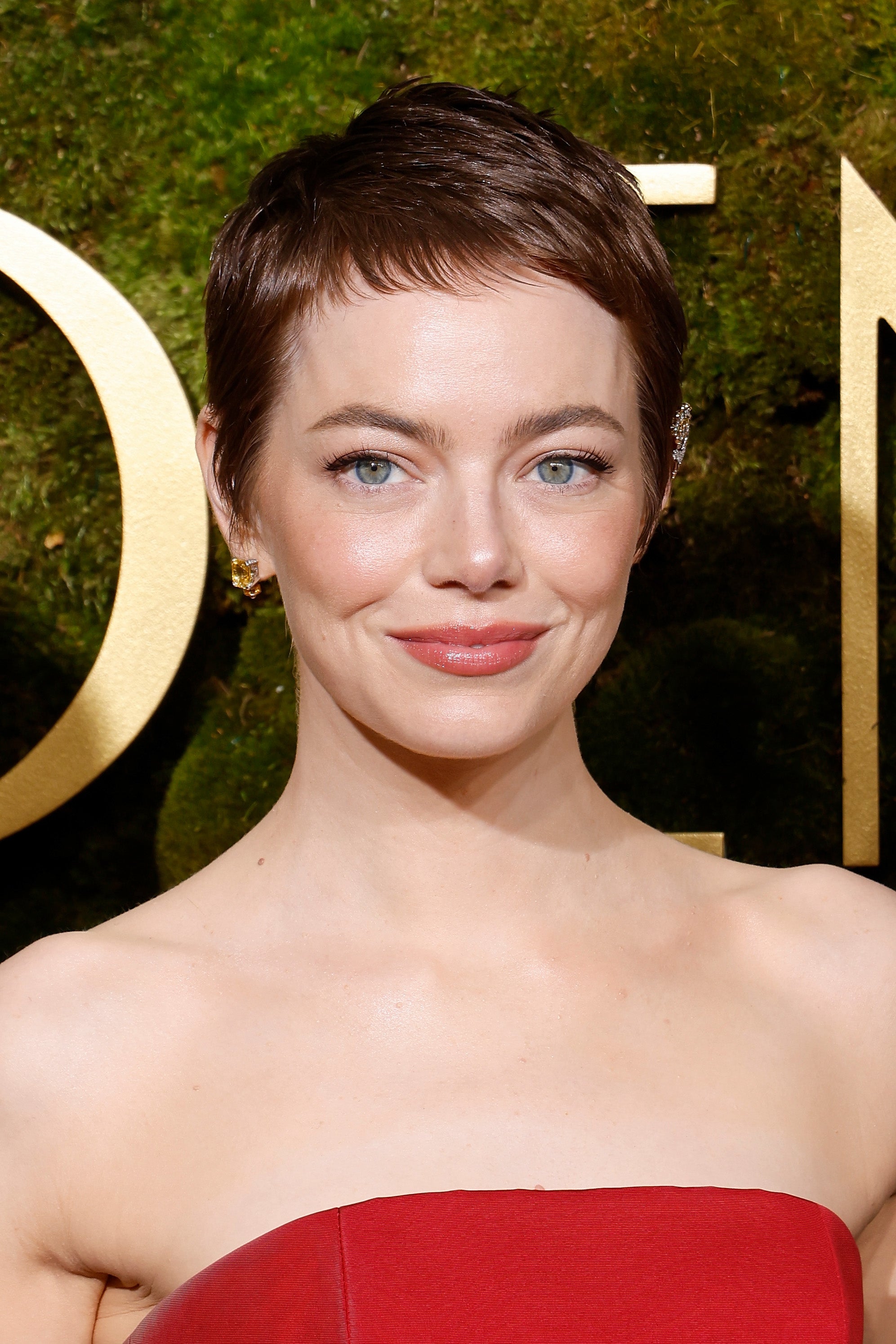 Person with short hair smiling, wearing an elegant strapless dress, earrings visible. Background has a textured, natural appearance