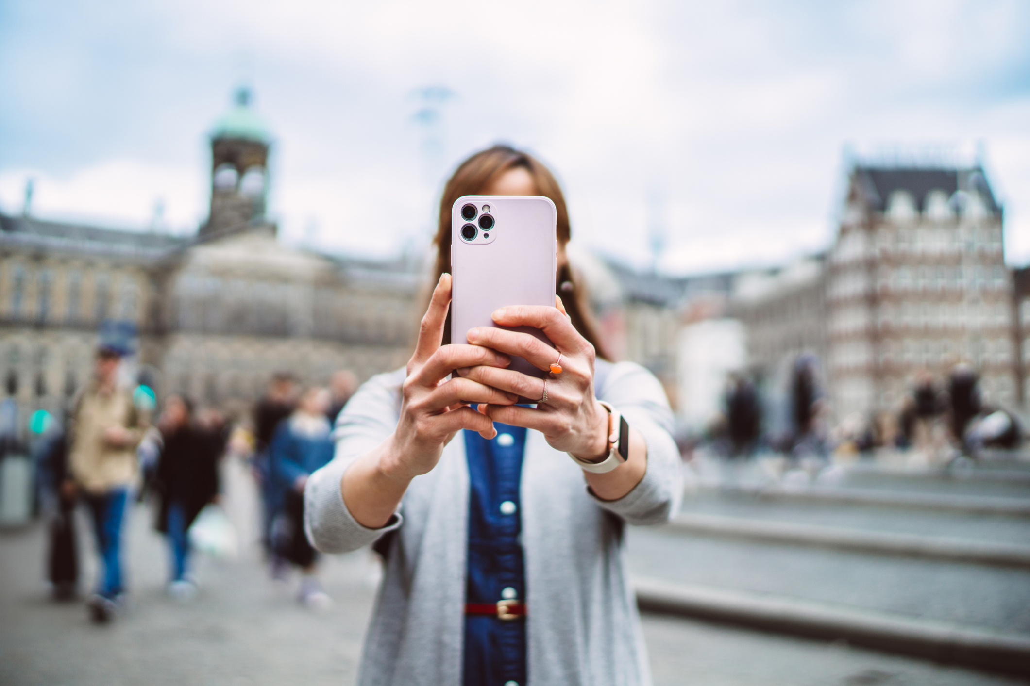 Person i uformell antrekk som tar en selfie med en smarttelefon på et bytorg, uskarpe historiske bygninger i bakgrunnen