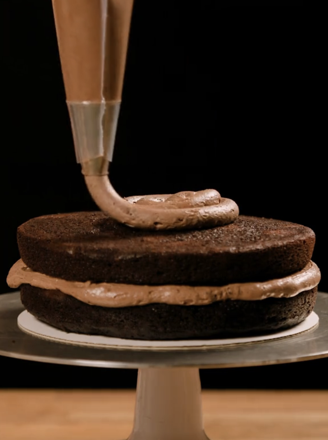 A close-up of a barroom   being decorated with cocoa  frosting from a piping bag, with 2  stacked layers of cocoa  cake