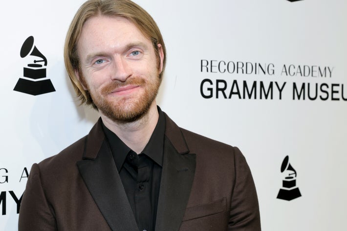 Finneas in a dark suit smiles at an event backdrop featuring the Recording Academy Grammy Museum logo