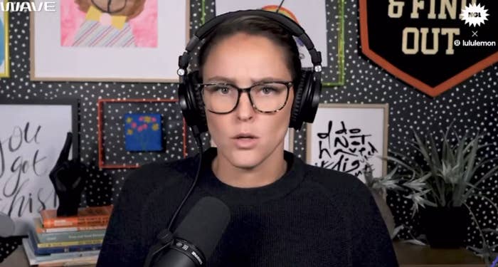 Kylie Kelce wearing headphones and glasses speaking into a microphone, with art and motivational posters in the background