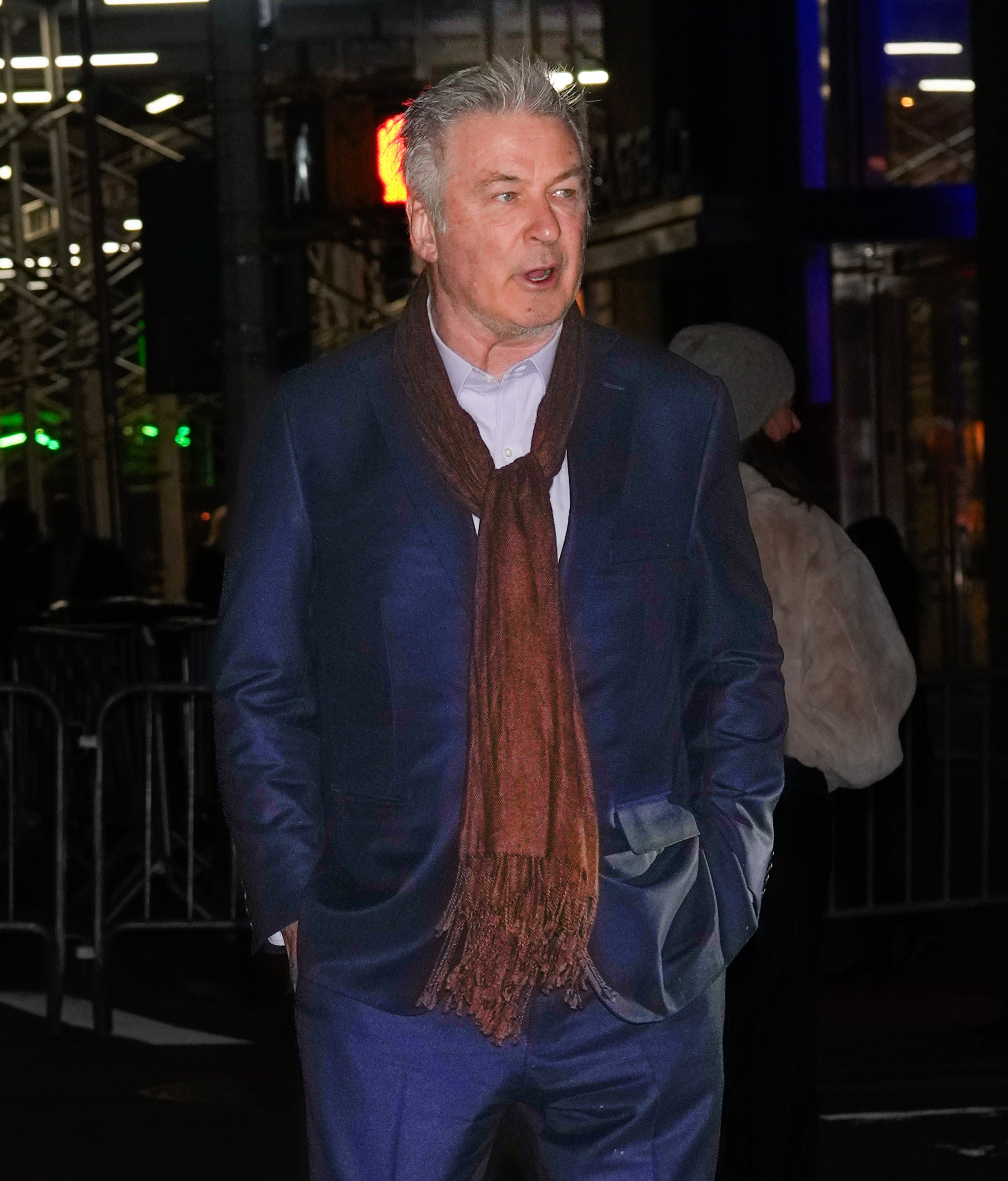 Person in a suit with a scarf, walking outside at night. Background shows lights and a building structure