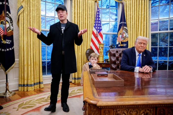Three people in the Oval Office: a man in a coat and cap gesturing, a child in casual attire, and another man seated at the desk in a suit