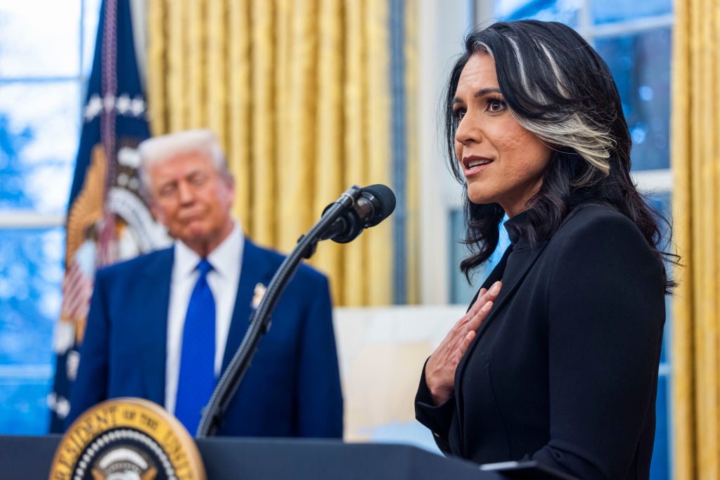 A woman speaks at a podium in formal attire while a man in a suit stands in the background in a room with golden curtains