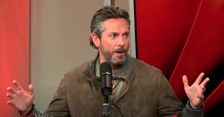 A man sitting in front of a microphone gestures with both hands during an interview