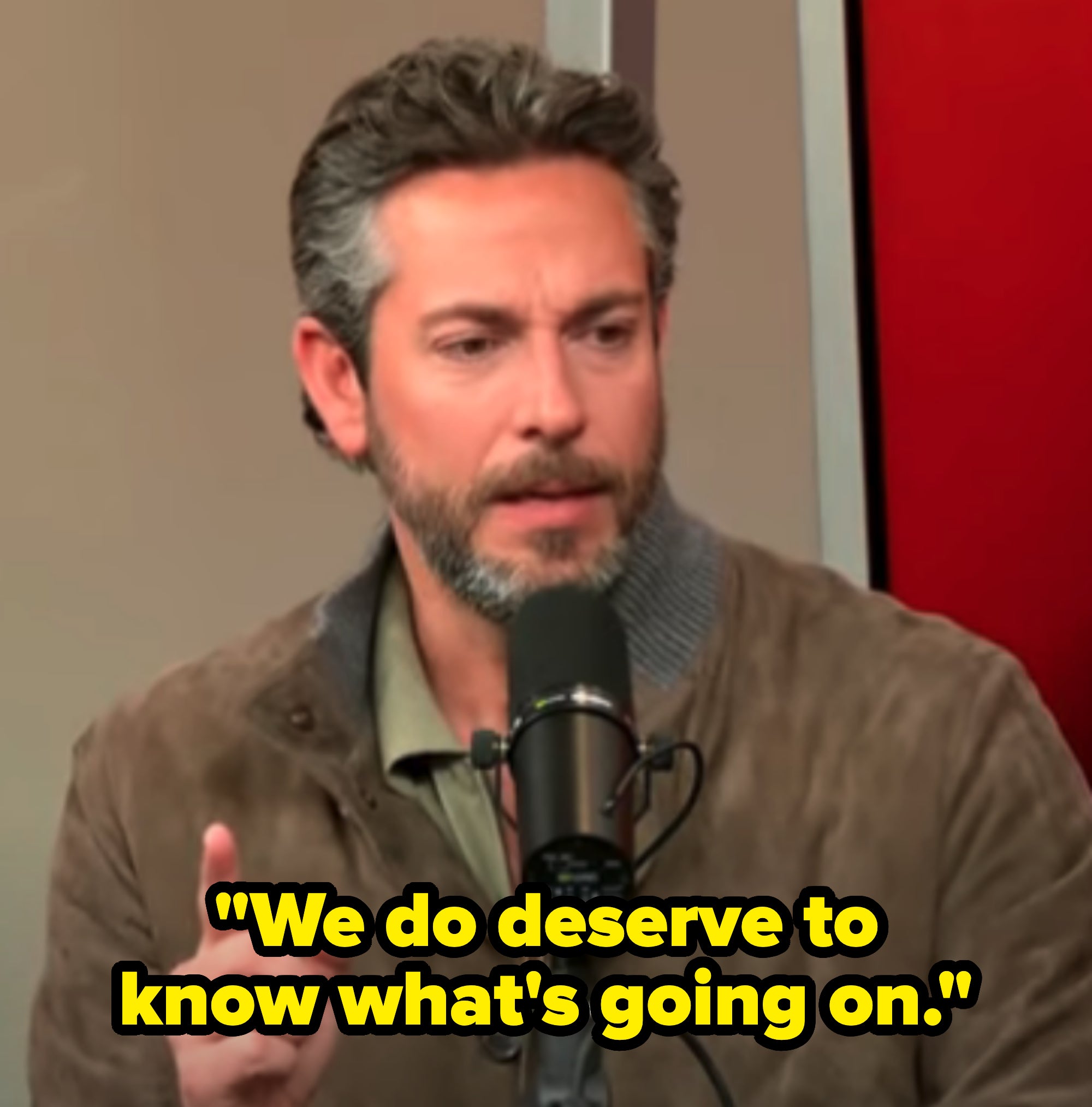 A man with short hair and a beard speaks into a microphone during an interview, gesturing with his hand