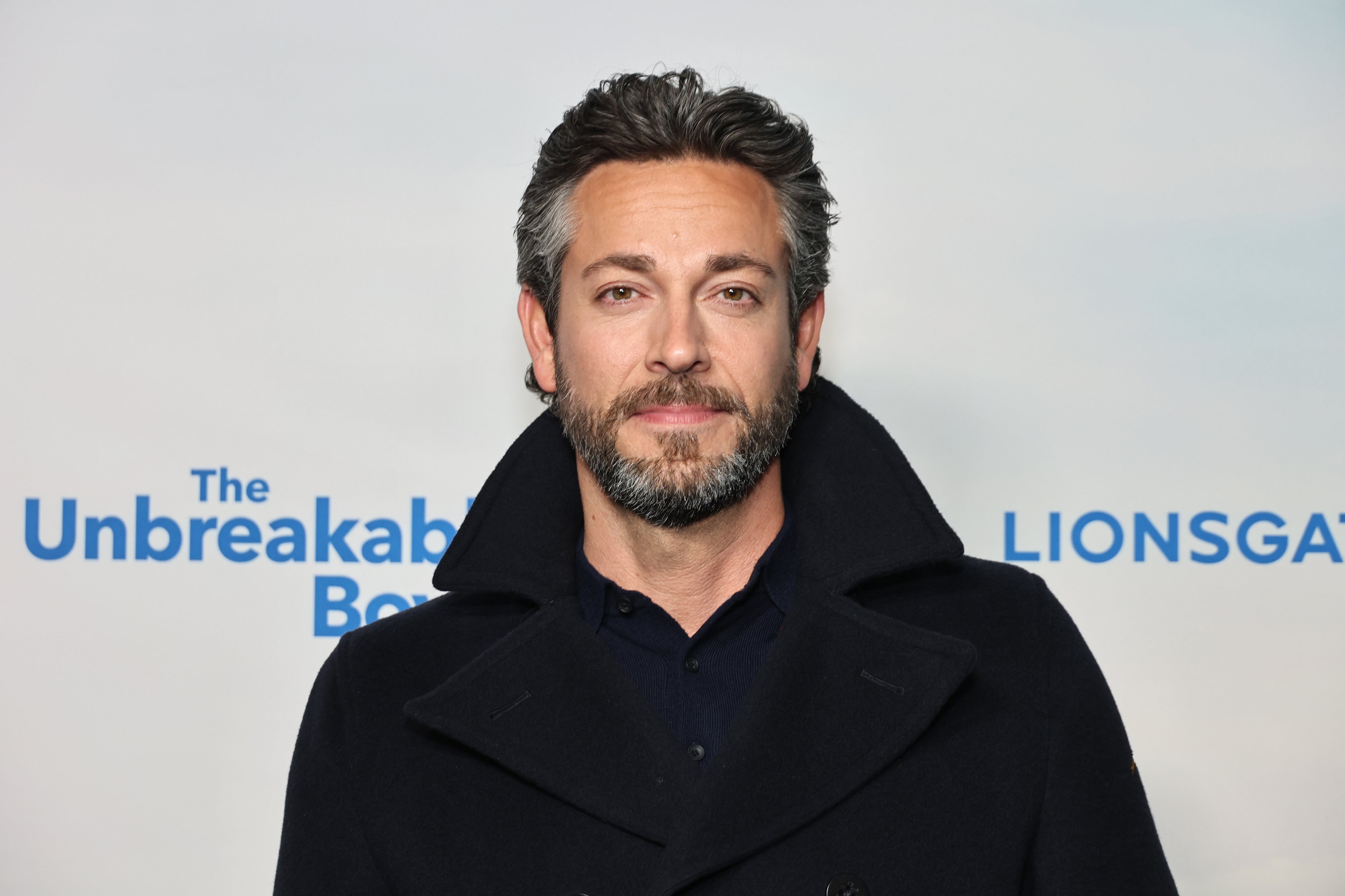 Man wearing a dark coat, standing in front of a promotional backdrop for "The Unbreakable Boy," at a media event