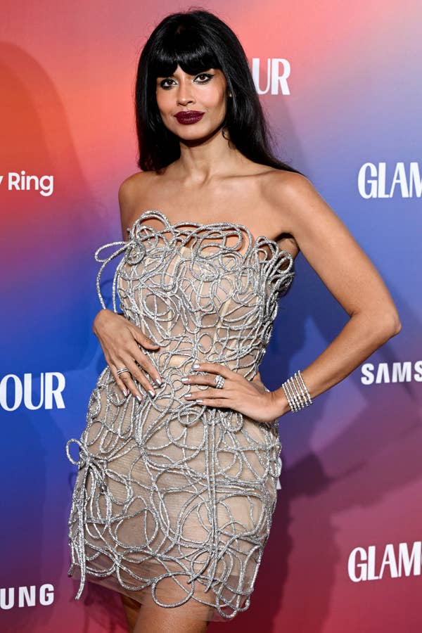 Person in an intricate, looping dress on a red carpet at a Glamour event, posing confidently with bold makeup