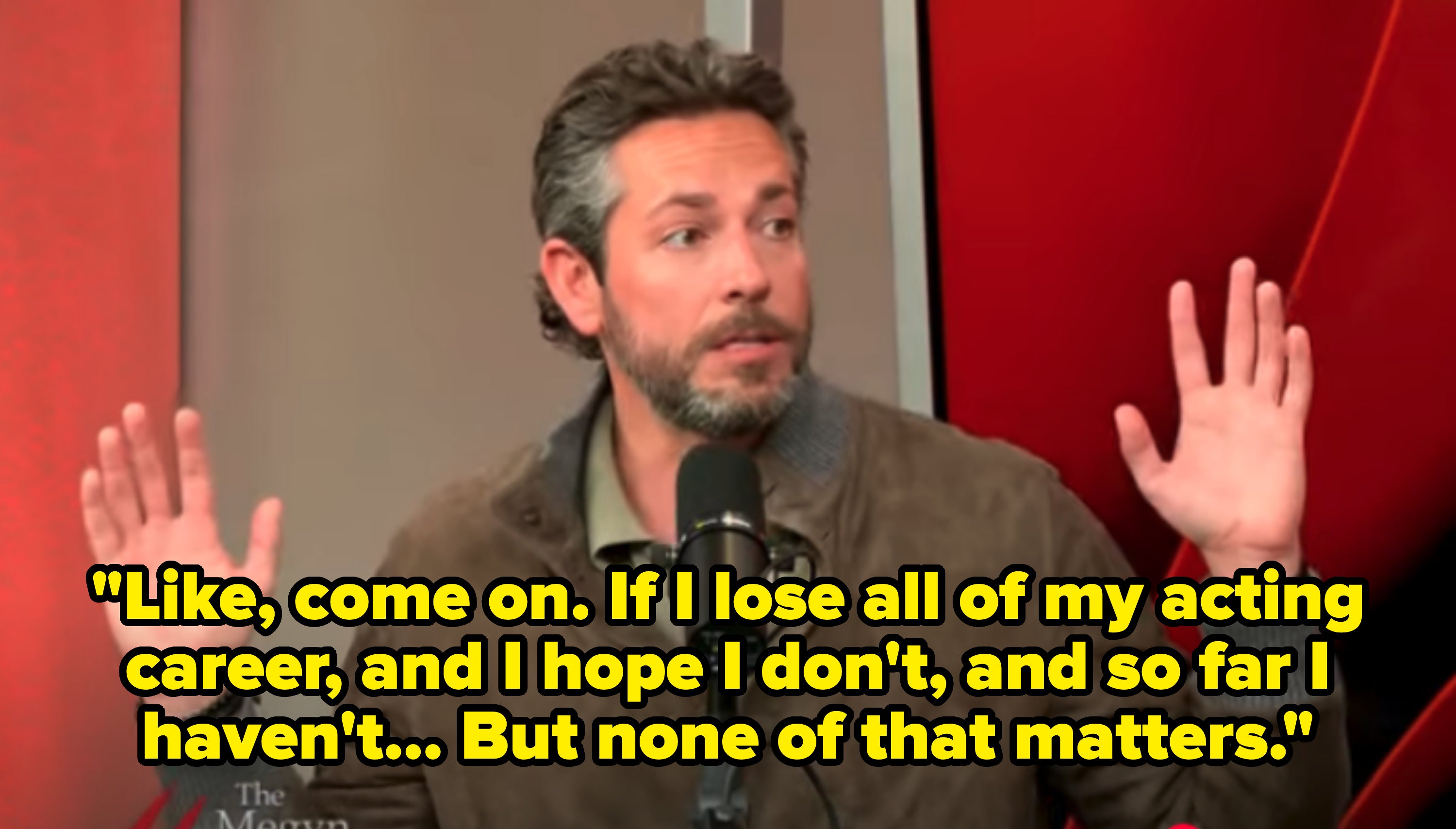 Man in suede jacket raises hands during a podcast interview, appearing thoughtful and engaged in conversation