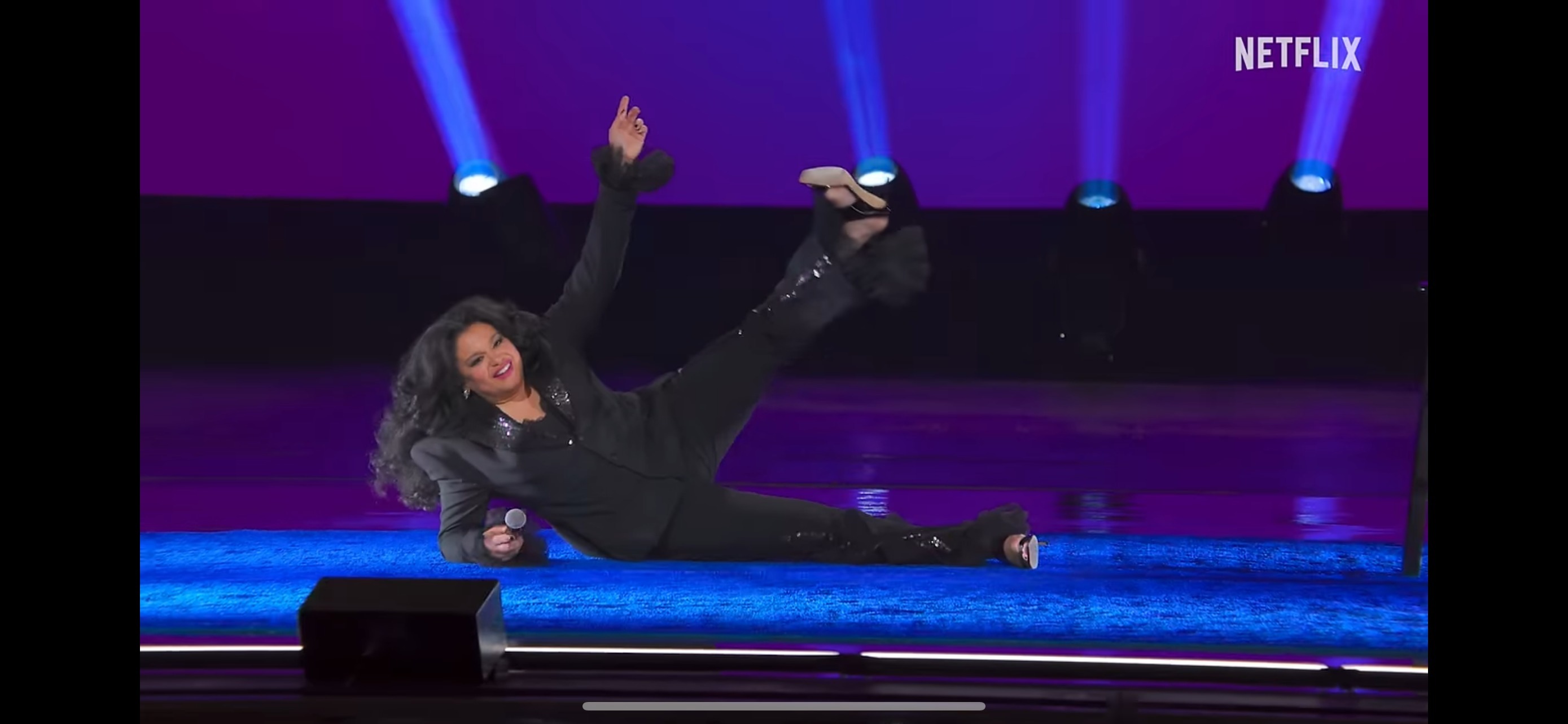 A person performs energetically on stage, lying down with one leg raised, holding a microphone, wearing a stylish black outfit