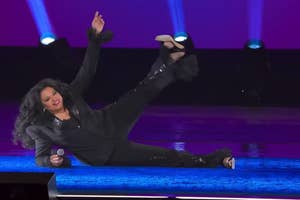 A person performs energetically on stage, lying down with one leg raised, holding a microphone, wearing a stylish black outfit