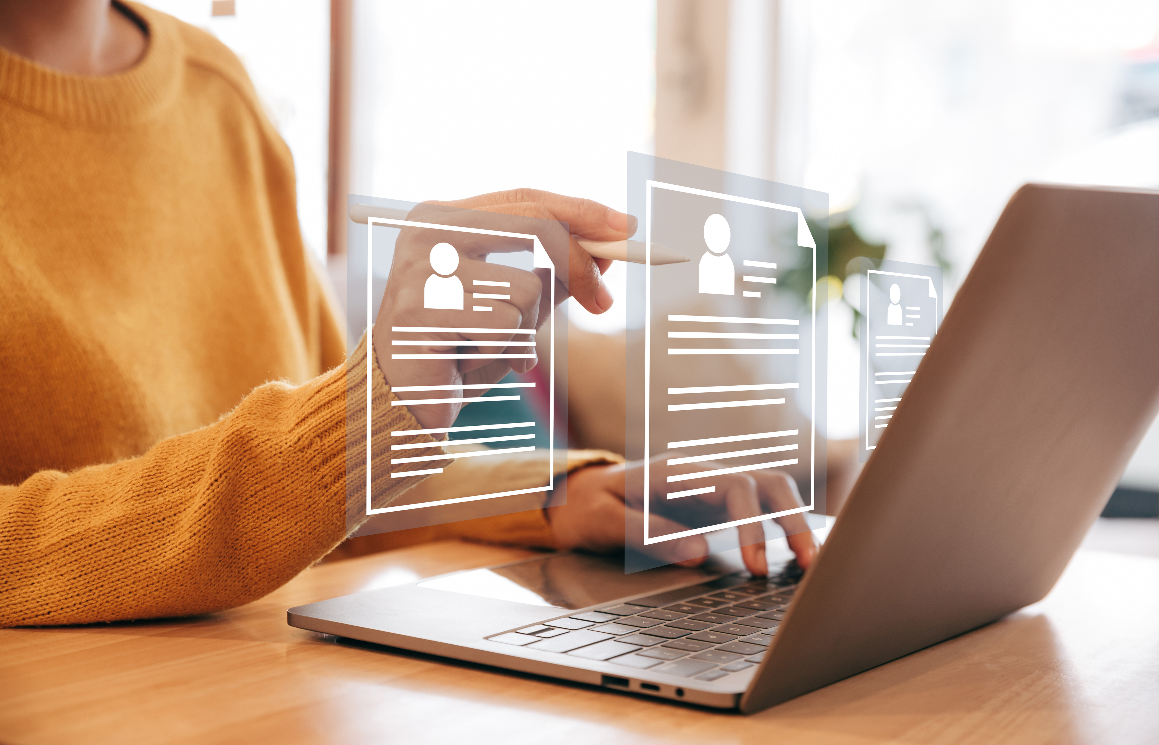Person in a sweater uses a laptop with floating holographic icons representing profiles, suggesting digital networking or job applications
