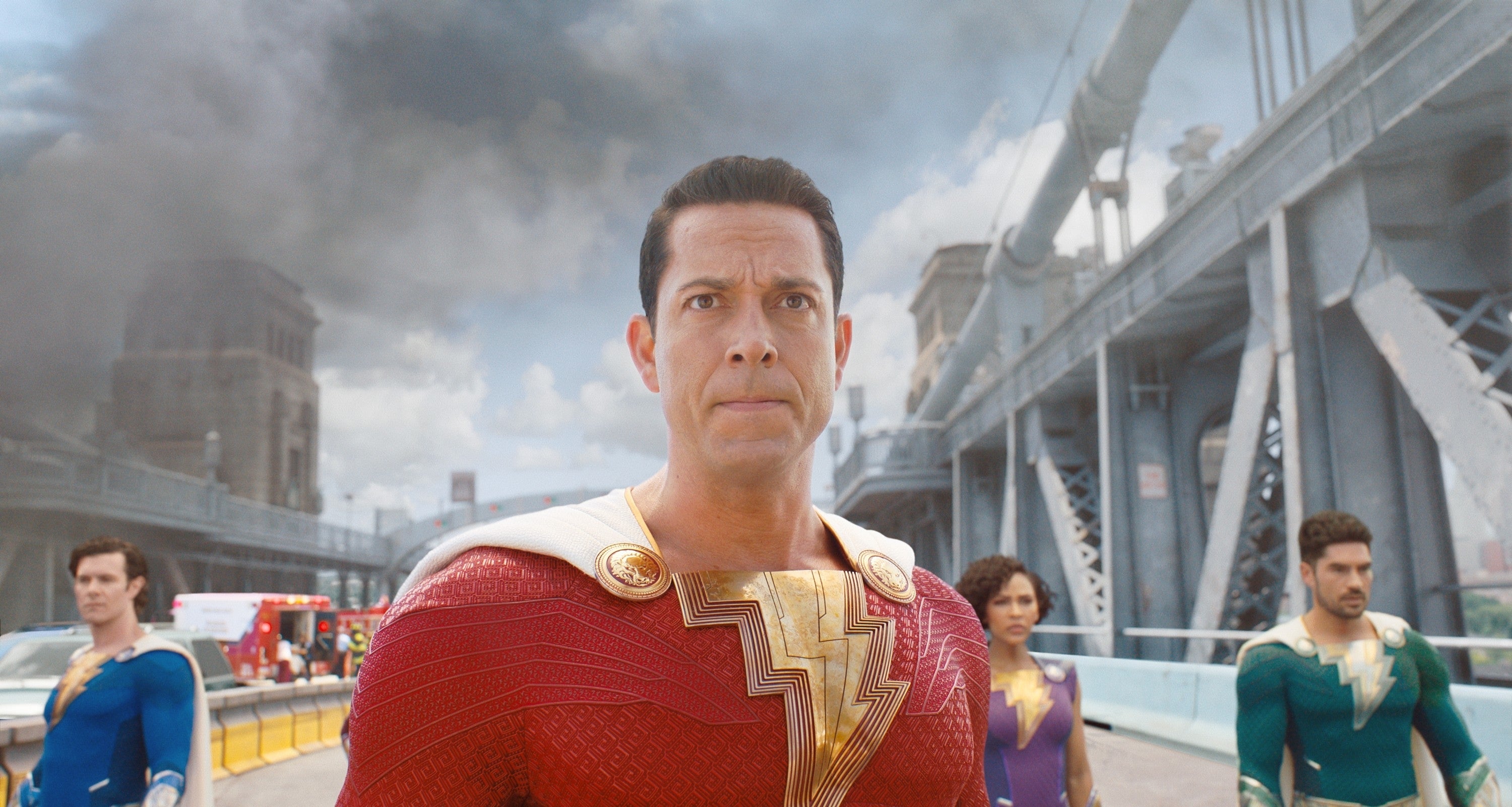 Actors in superhero costumes stand on a city bridge. The central figure wears a lightning bolt emblem. Smoke rises in the background
