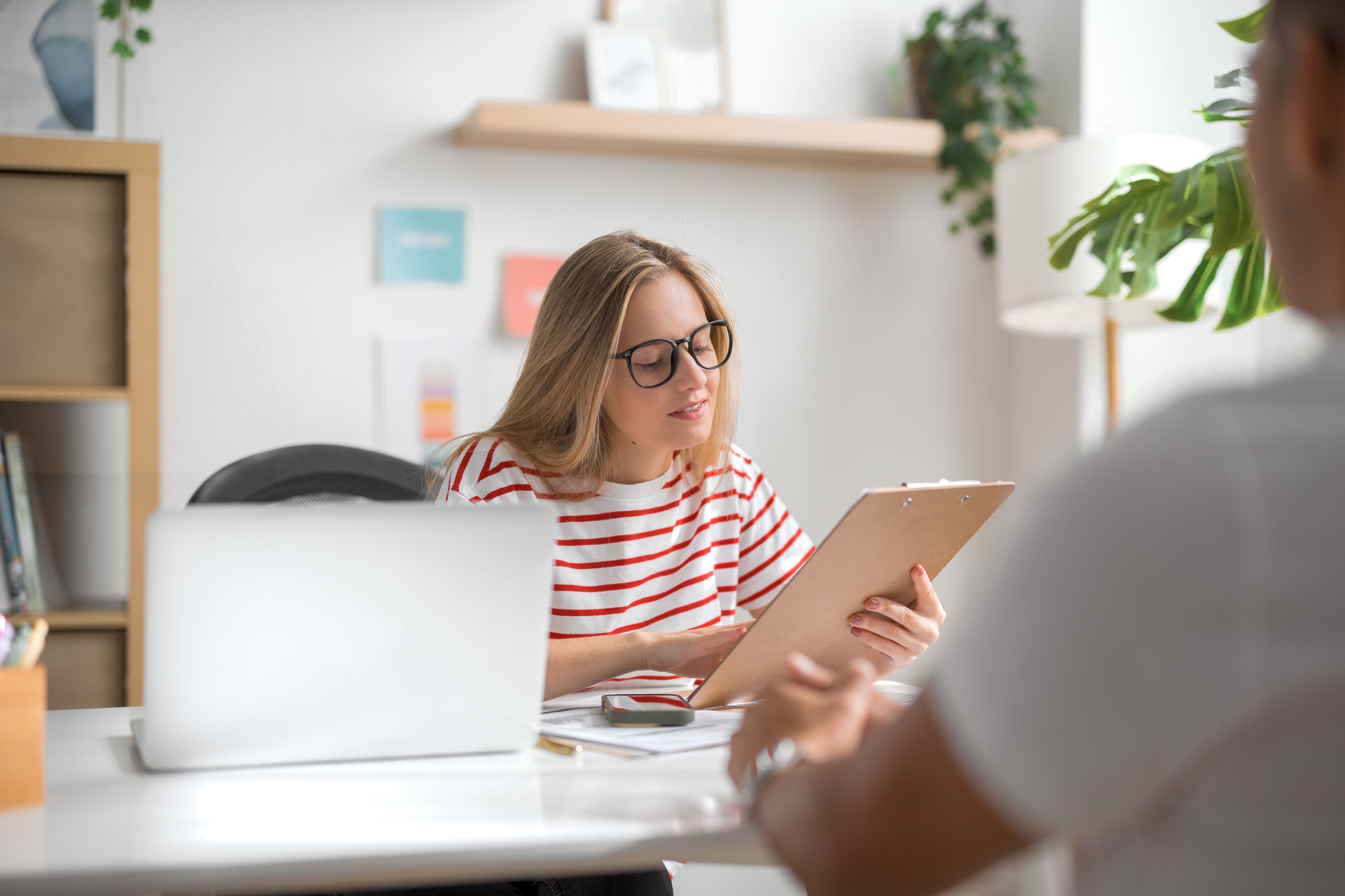 Woman in glasses and striped shirt holding clipboard, discussing with a person off camera in a modern work setting. Laptop and plants visible