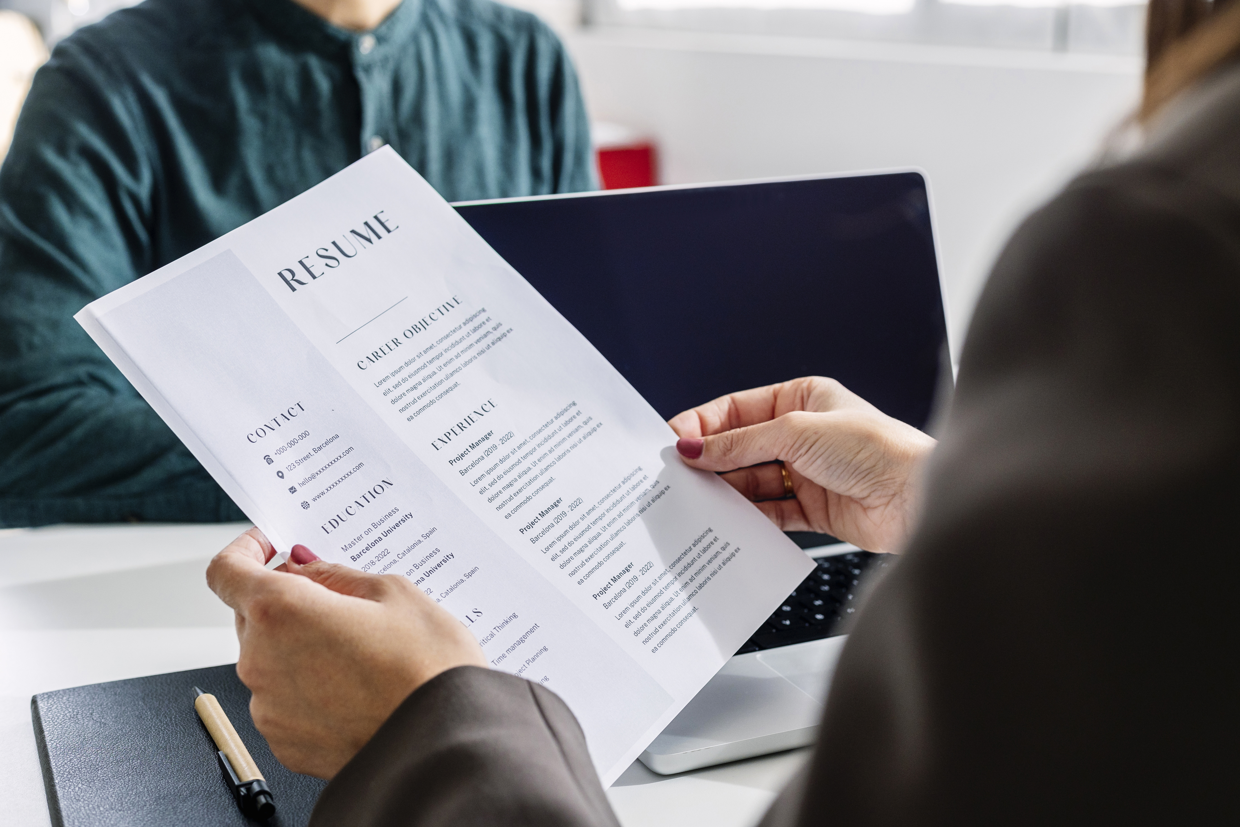 Person holding a resume in a professional setting, possibly during a job interview. Another person is visible, sitting near a laptop