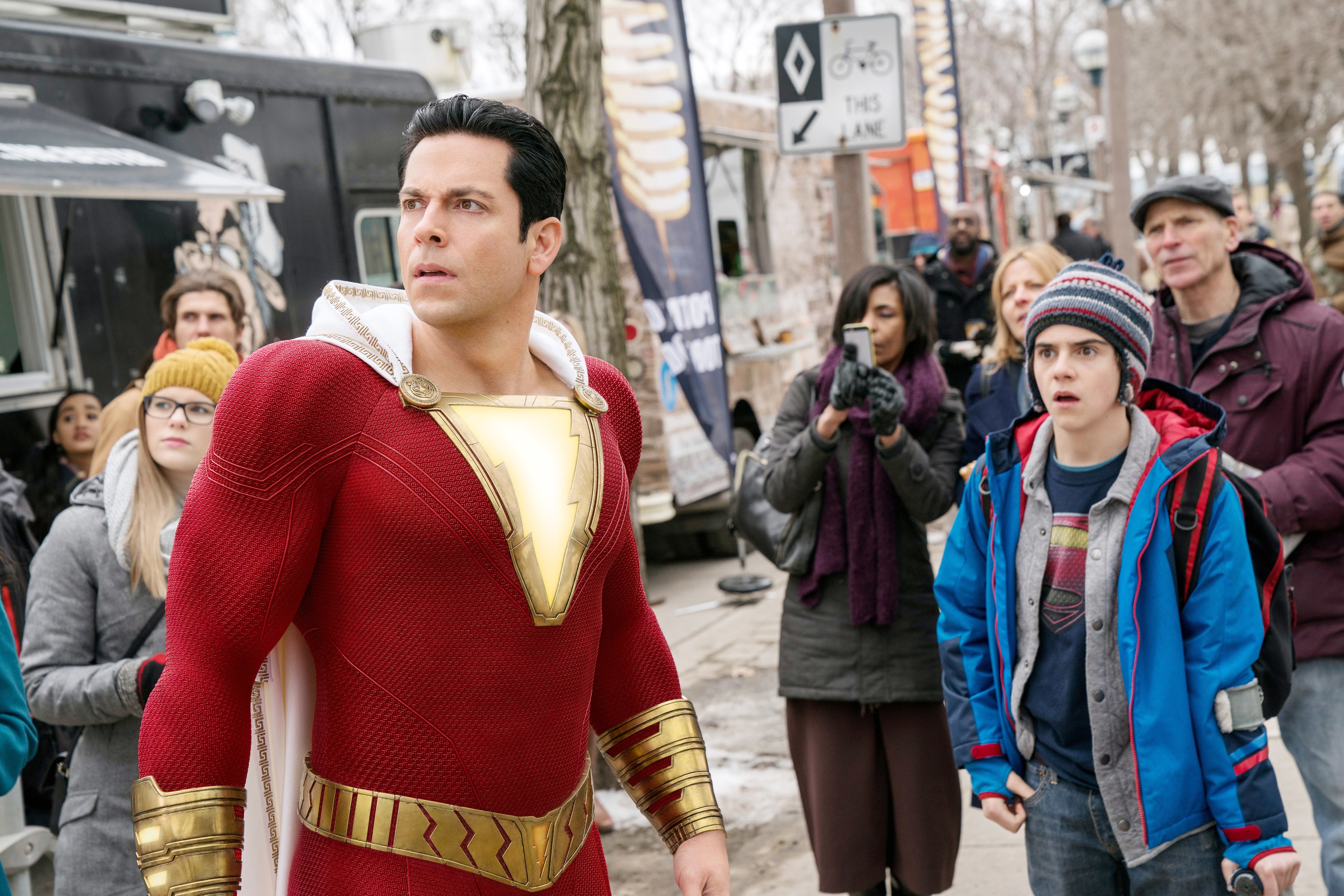 Actor in superhero costume stands on crowded street; onlookers appear surprised and intrigued