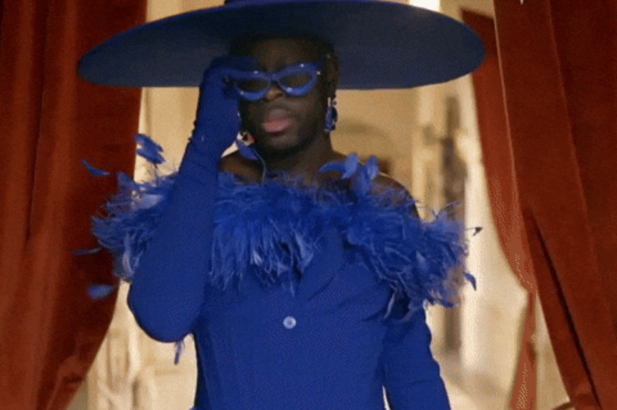 Person in glamorous, large hat and feathered outfit on a runway, adjusting vibrant glasses in stylish manner