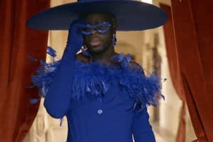 Person in glamorous, large hat and feathered outfit on a runway, adjusting vibrant glasses in stylish manner