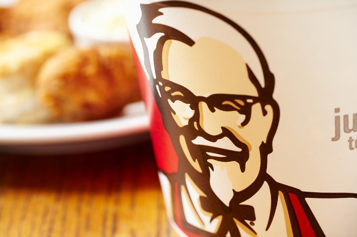 カーネル・サンダースのイラストが描かれたKFCのチキンバケットがテーブルに置かれている。バックにチキン料理が見える。