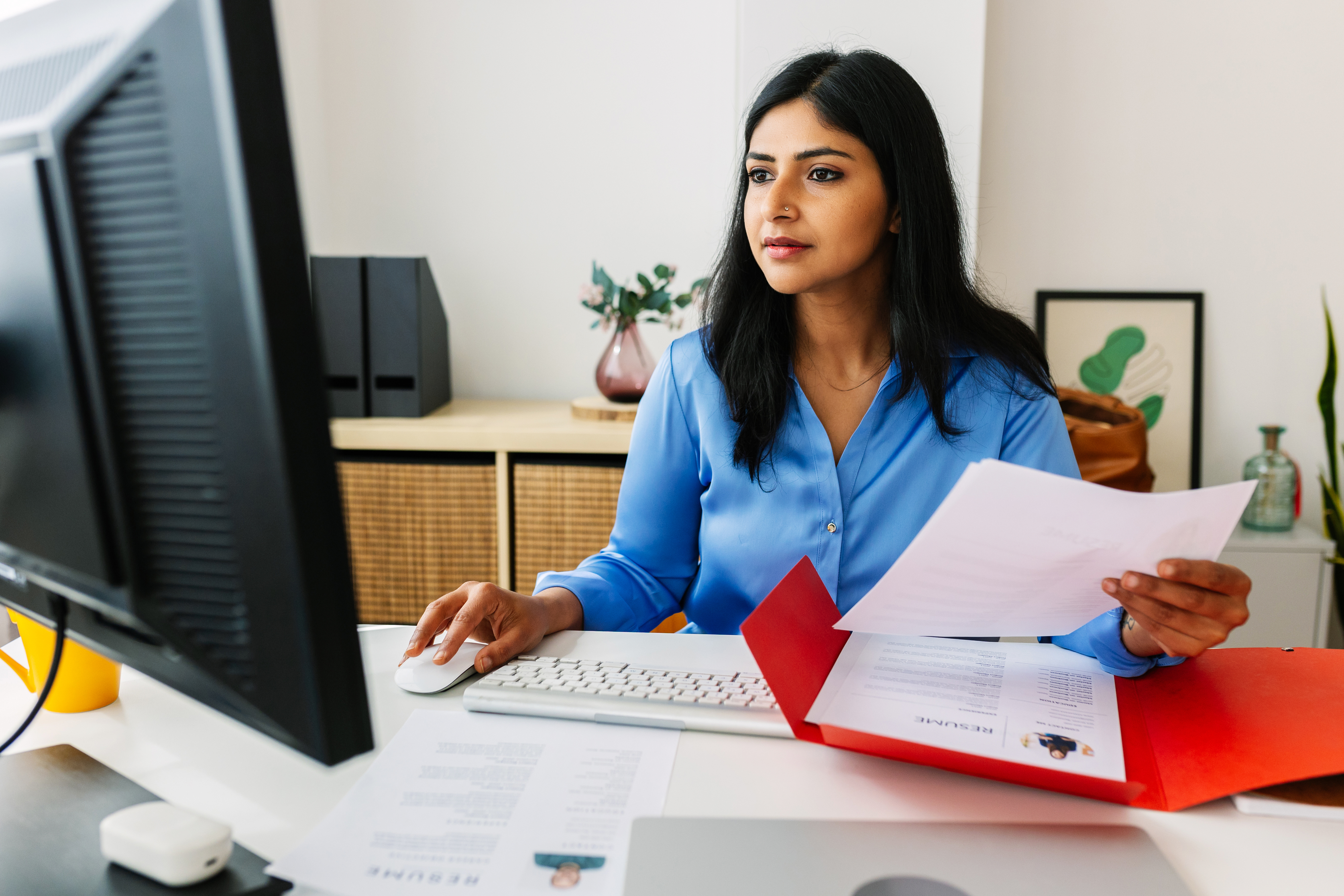 Person in a smart casual outfit works at a computer, holding documents and focusing on the screen in an office setting