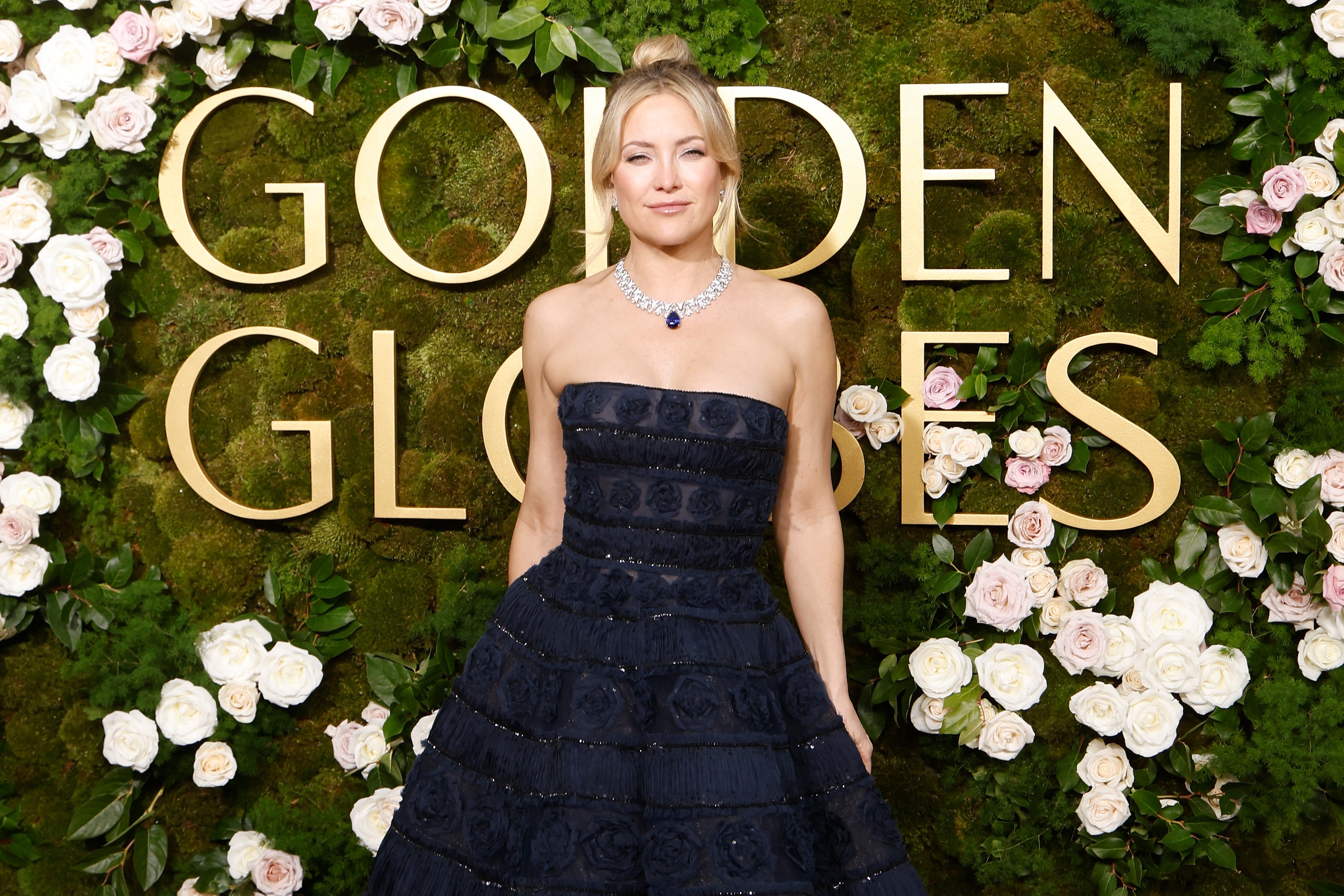 Kate Hudson in strapless gown stands before "Golden Globes" backdrop, surrounded by flowers