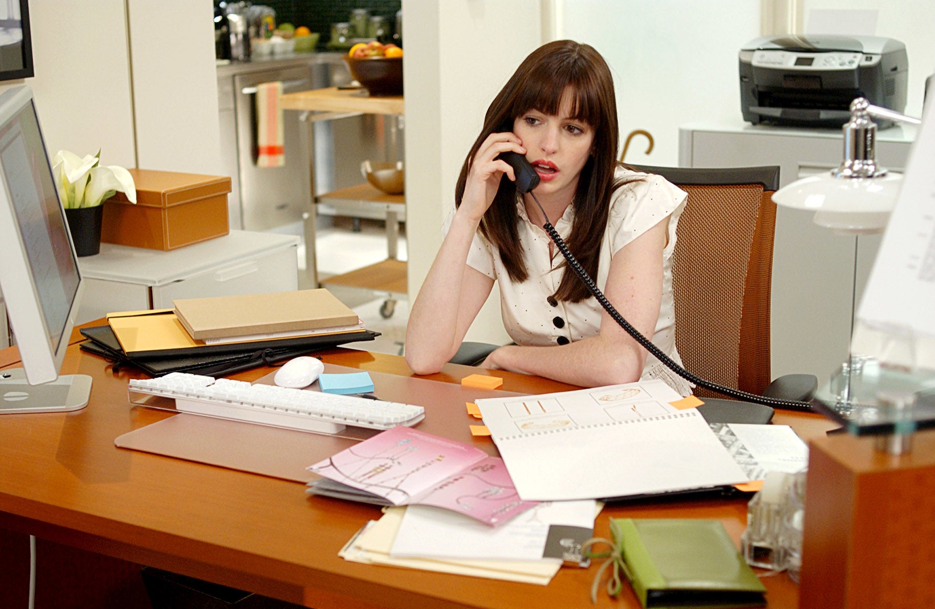 Anne Hathaway sitting at an office desk, talking on the phone, surrounded by papers and office supplies