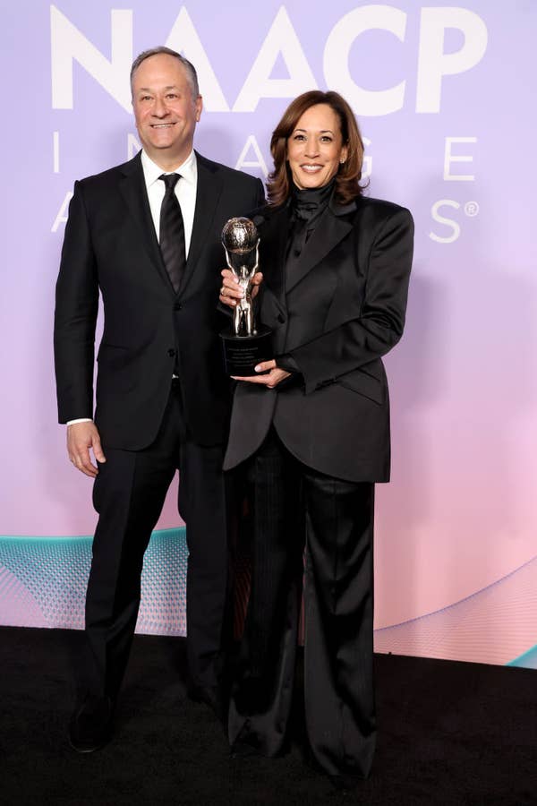 Two people on a red carpet, one holding an award. Both wear formal black attire, including a suit and a tuxedo. NAACP Image Awards backdrop