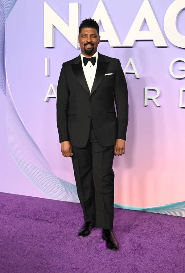 Person in a black tuxedo with a bow tie standing on a purple carpet at a celebrity event