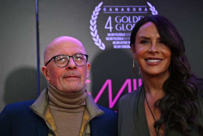 Two people posing on a red carpet. One wears glasses and a turtleneck under a jacket, the other wears a deep V-neck dress with hoop earrings