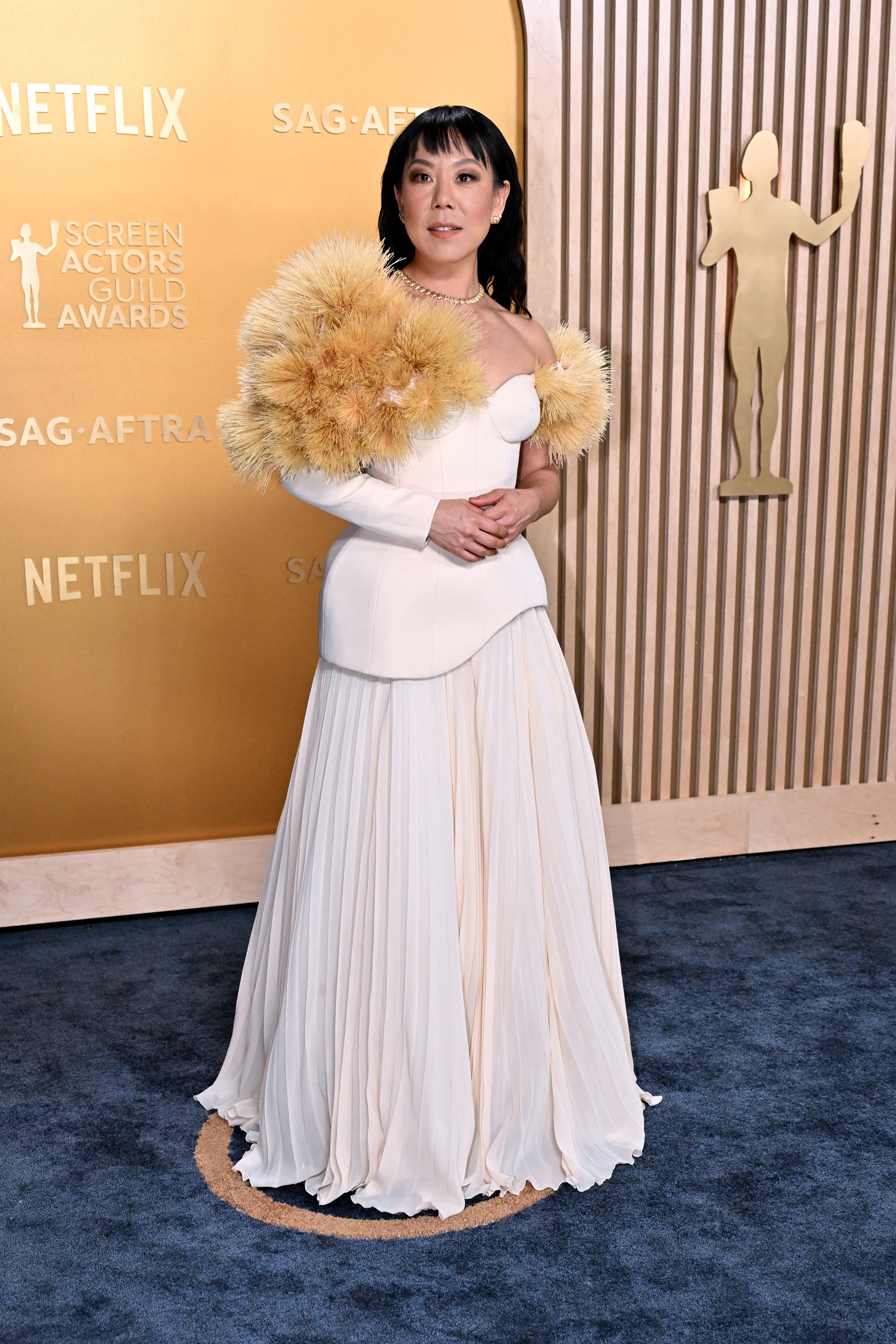 Person in elegant white gown with fluffy shoulder details at an awards event, standing on a blue carpet backdrop with event logos