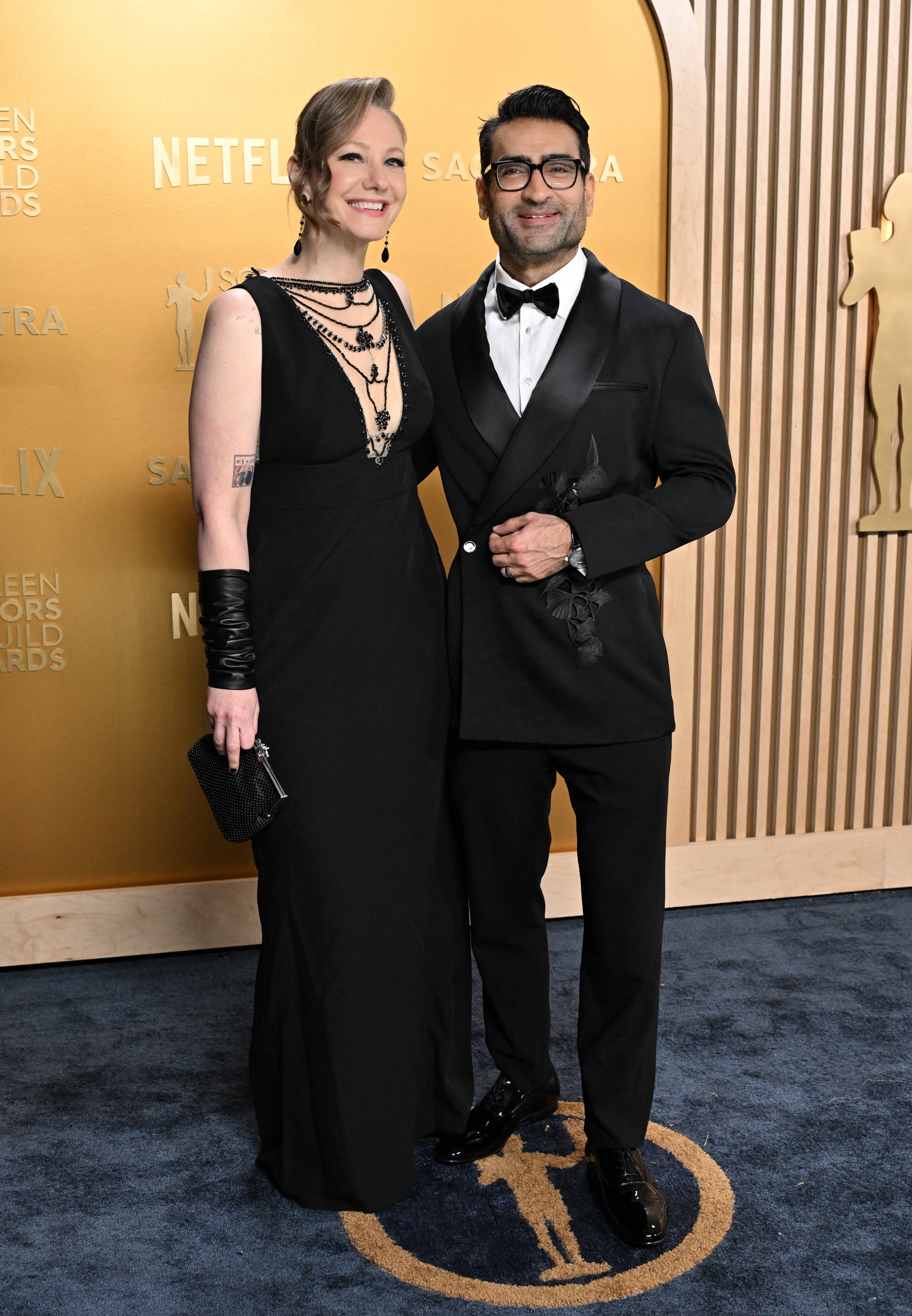 Two people pose on a red carpet. One wears a black sleeveless gown and gloves; the other wears a black tuxedo with a bow tie