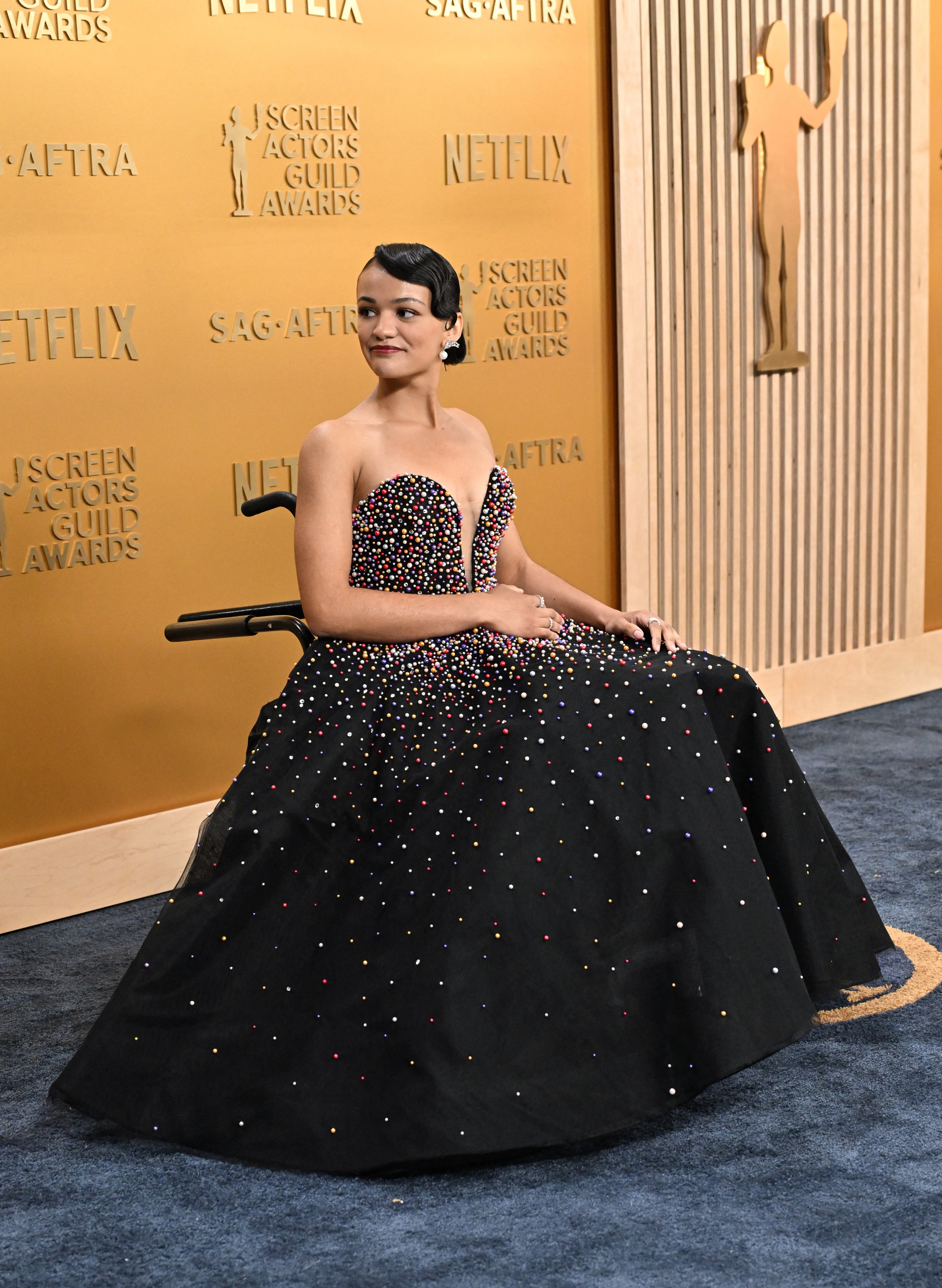 Person in a wheelchair wearing an elegant, beaded gown on a red carpet at a Screen Actors Guild Awards event